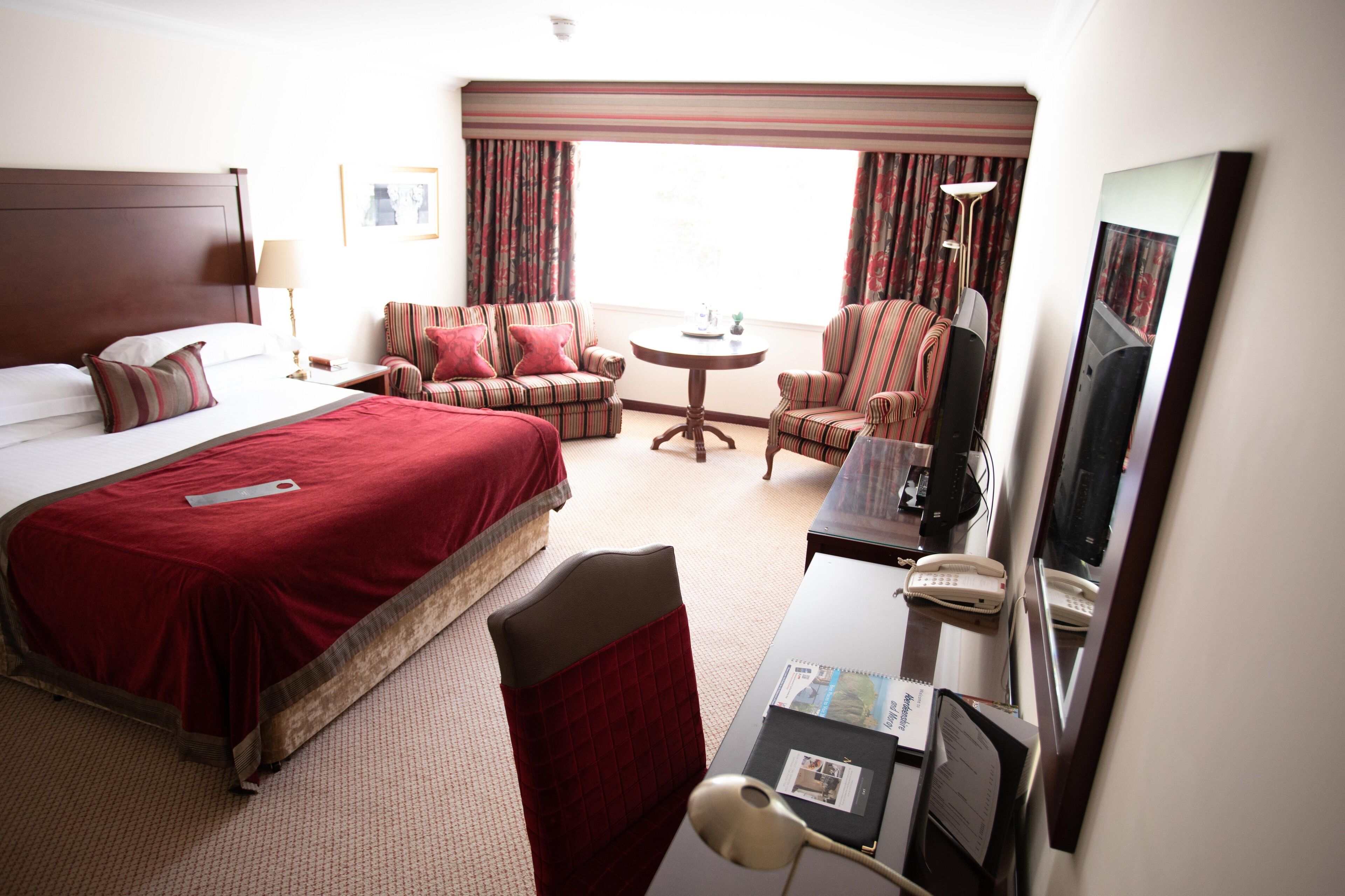 A hotel room with a large bed, two armchairs, a small round table, a desk with a telephone, and a television.