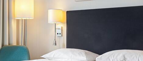A hotel room with a bed, two bedside lamps, a teal chair, and a framed quote on the wall.