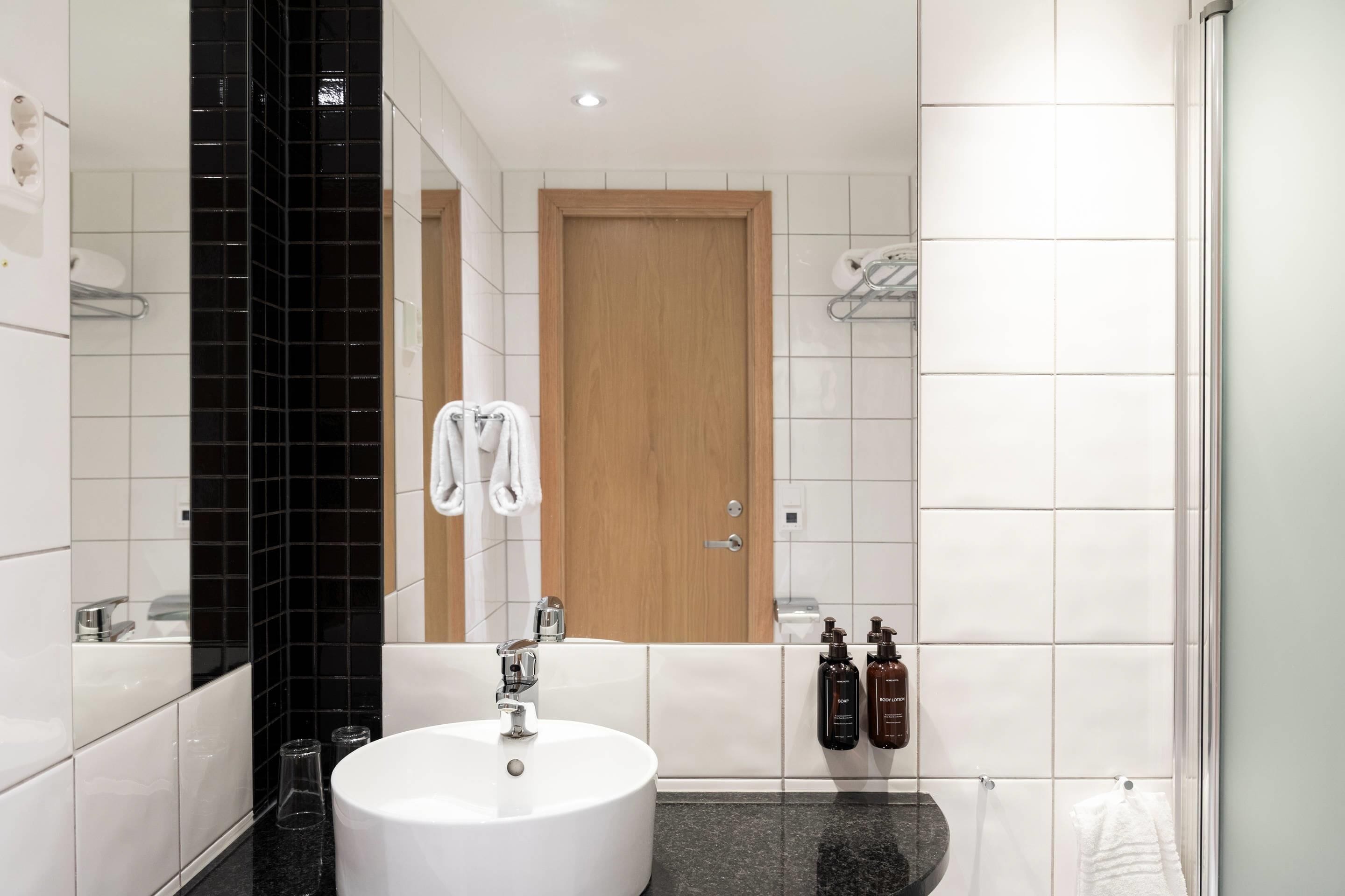 A modern bathroom with a white sink, black tiled backsplash, a wooden door, and two bottles of shampoo.