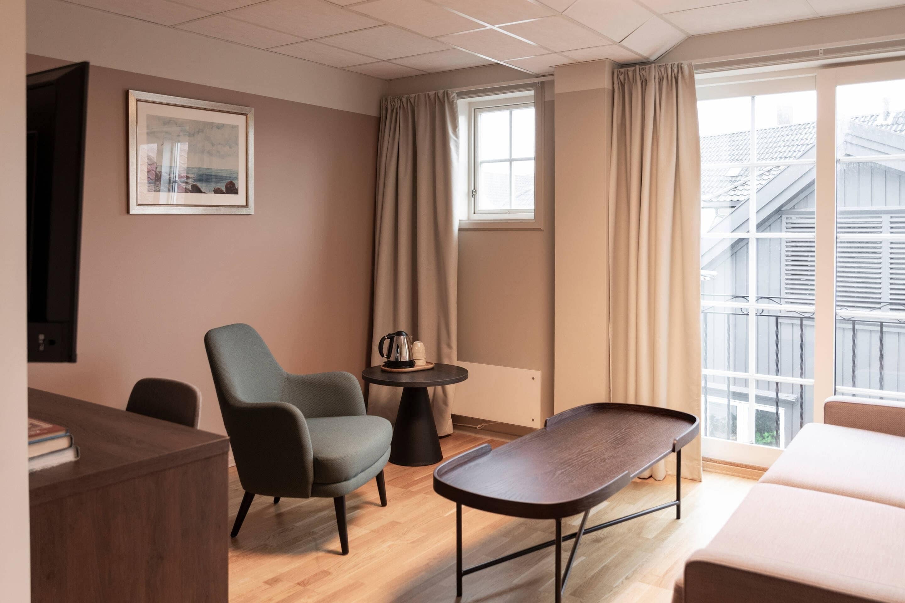 A modern hotel room with a wooden floor, a desk, a chair, a small table with a kettle, and a bed.