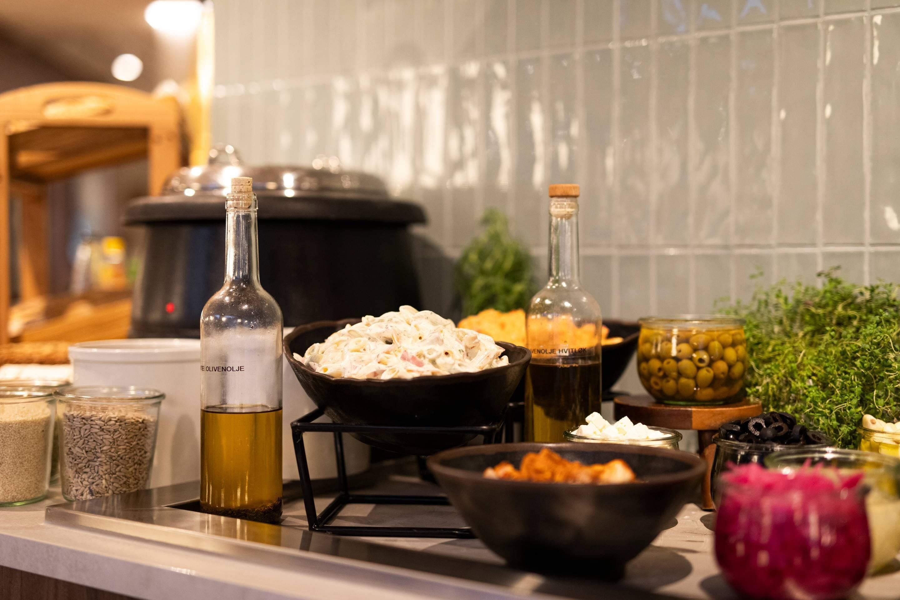 Una encimera de cocina con diversos alimentos y recipientes, incluyendo una botella de aceite de oliva, tazones de comida y frascos de aceitunas y encurtidos.