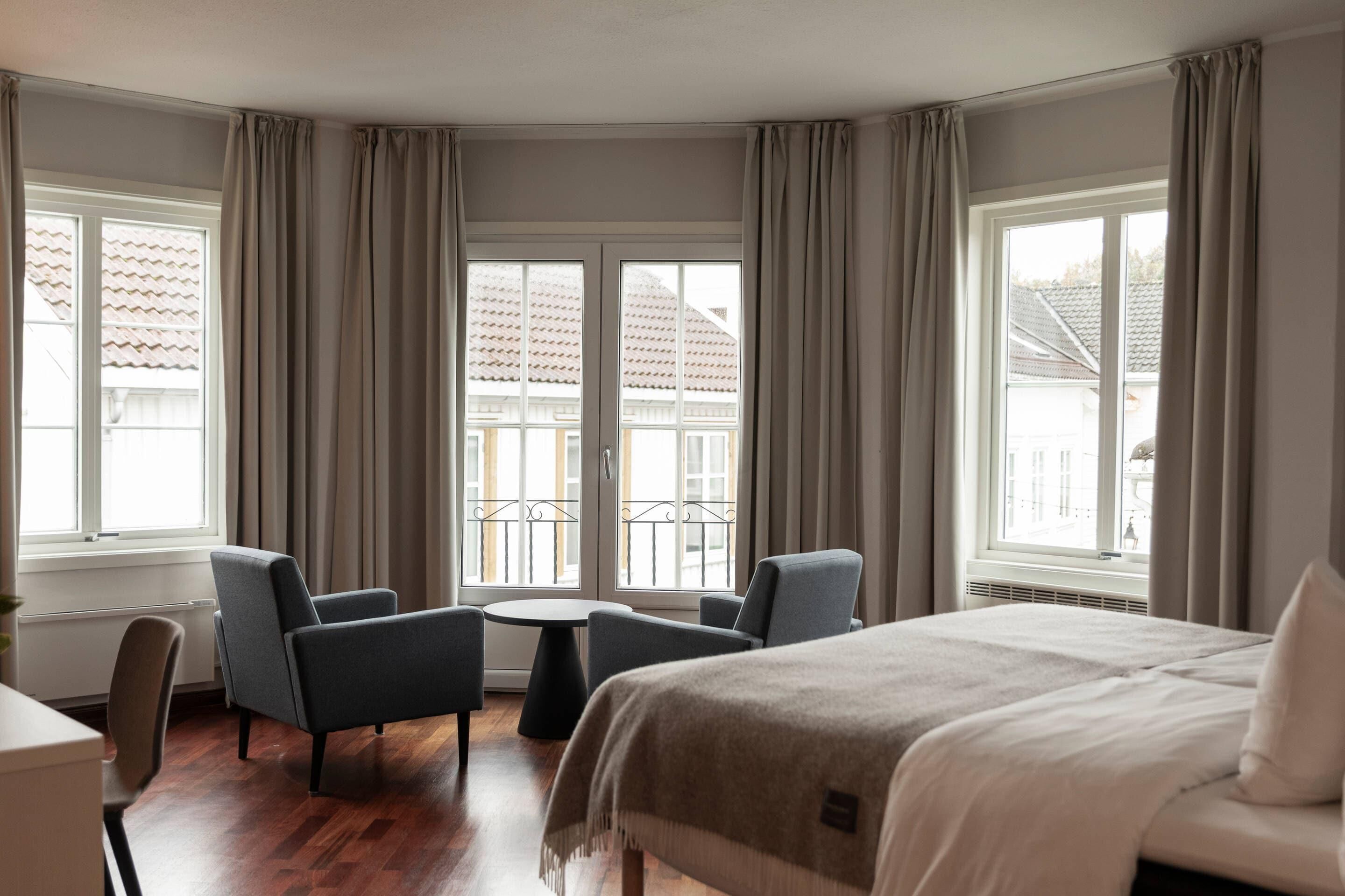 Una habitación de hotel con una cama, dos sillones, una mesita y ventanas grandes con cortinas.