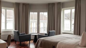 Una habitación de hotel con una cama, dos sillones, una mesita y ventanas grandes con cortinas.