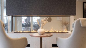 A modern interior with a fireplace, two upholstered chairs, and a large window overlooking a dining area. - Home Hotel Grimstad (Grimstad)