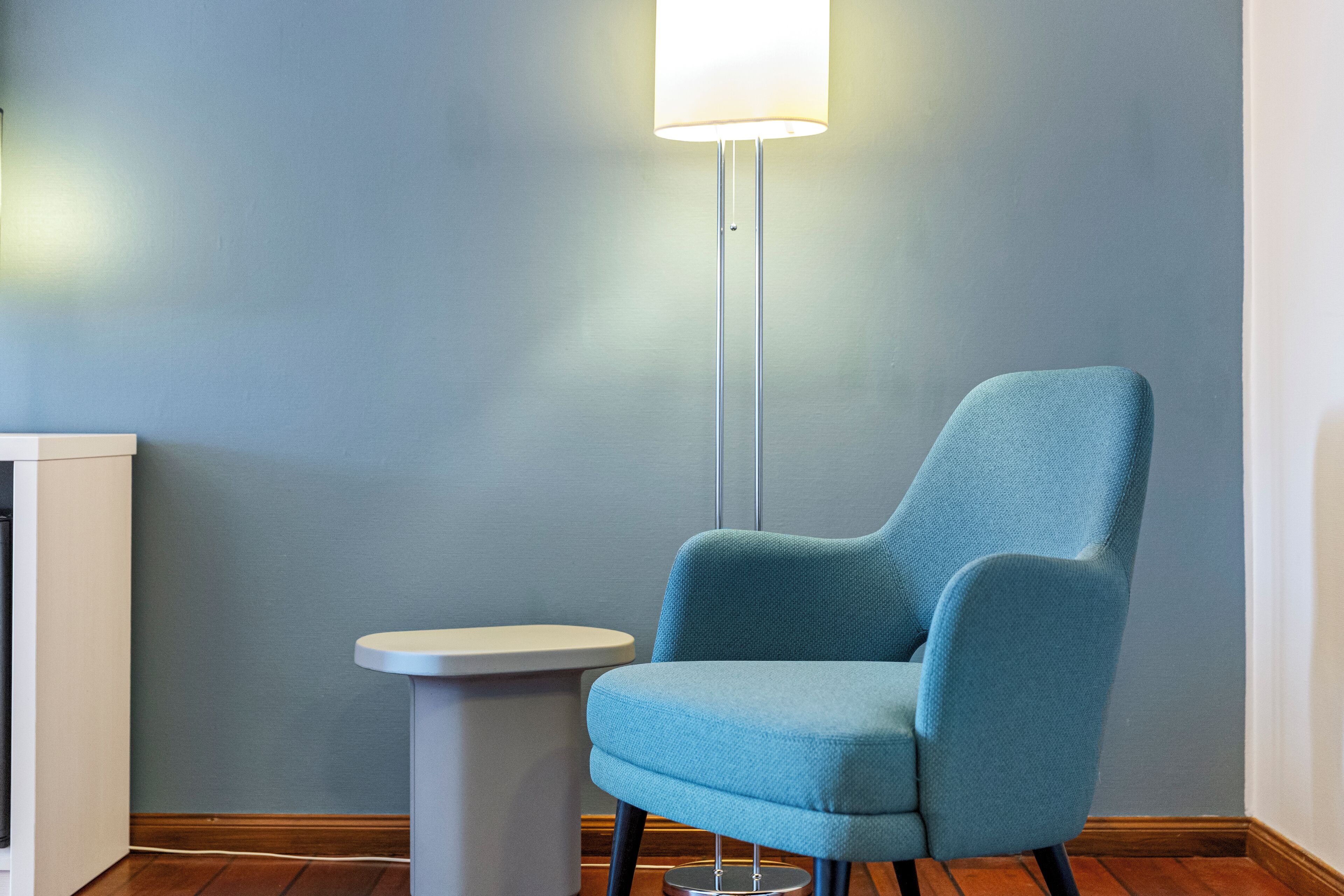 Una habitación con un sillón azul, una mesita blanca y una lámpara de pie frente a una pared azul.