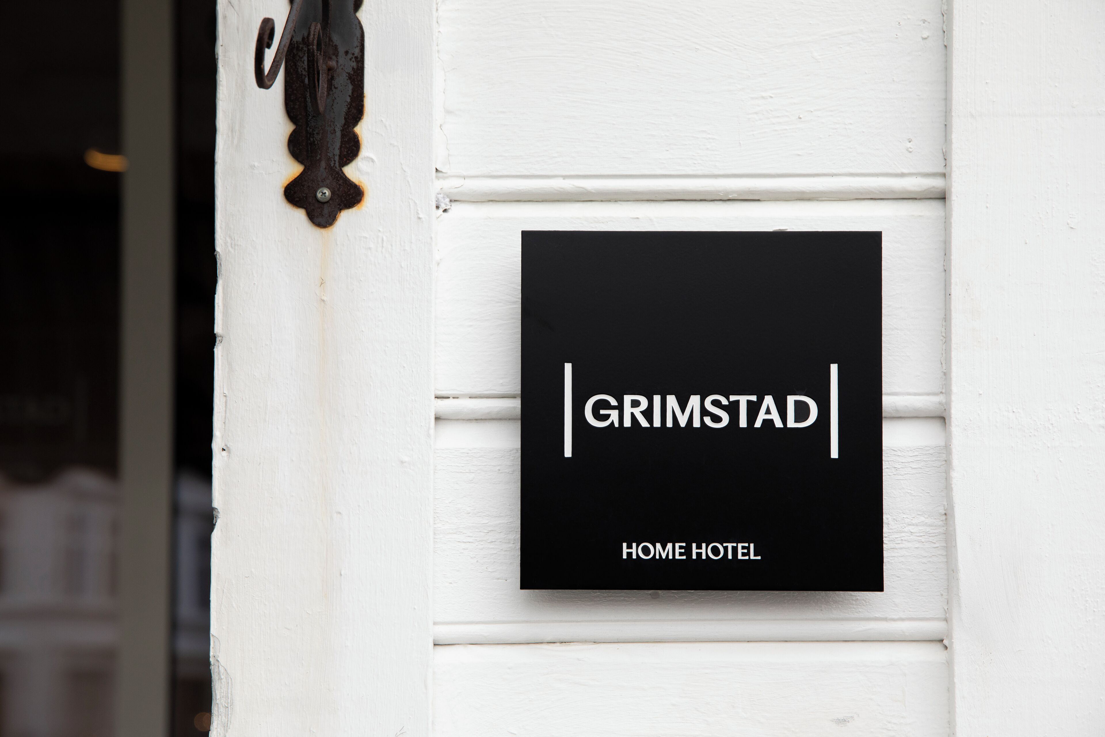 A black sign with white text reading "GRIMSTAD HOME HOTEL" mounted on a white wall.