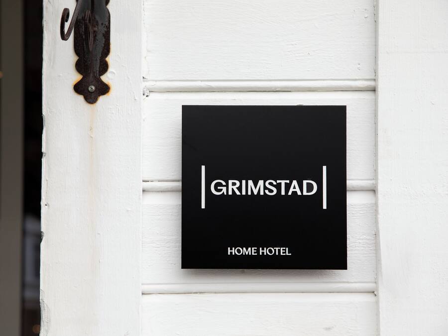 Un letrero negro con texto blanco que dice 'GRIMSTAD HOME HOTEL' montado sobre una pared blanca.