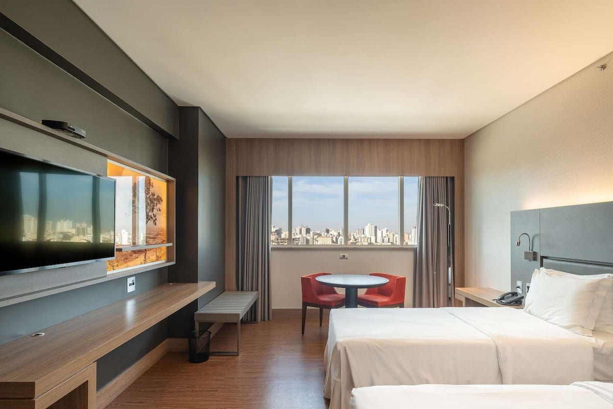 Une chambre dâhĂŽtel avec un lit, un bureau avec une tĂ©lĂ©vision, une petite table avec des chaises et une vue sur la ville.