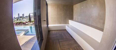A modern bathroom with a large bathtub, wooden flooring, and a view of a cityscape and pool area.