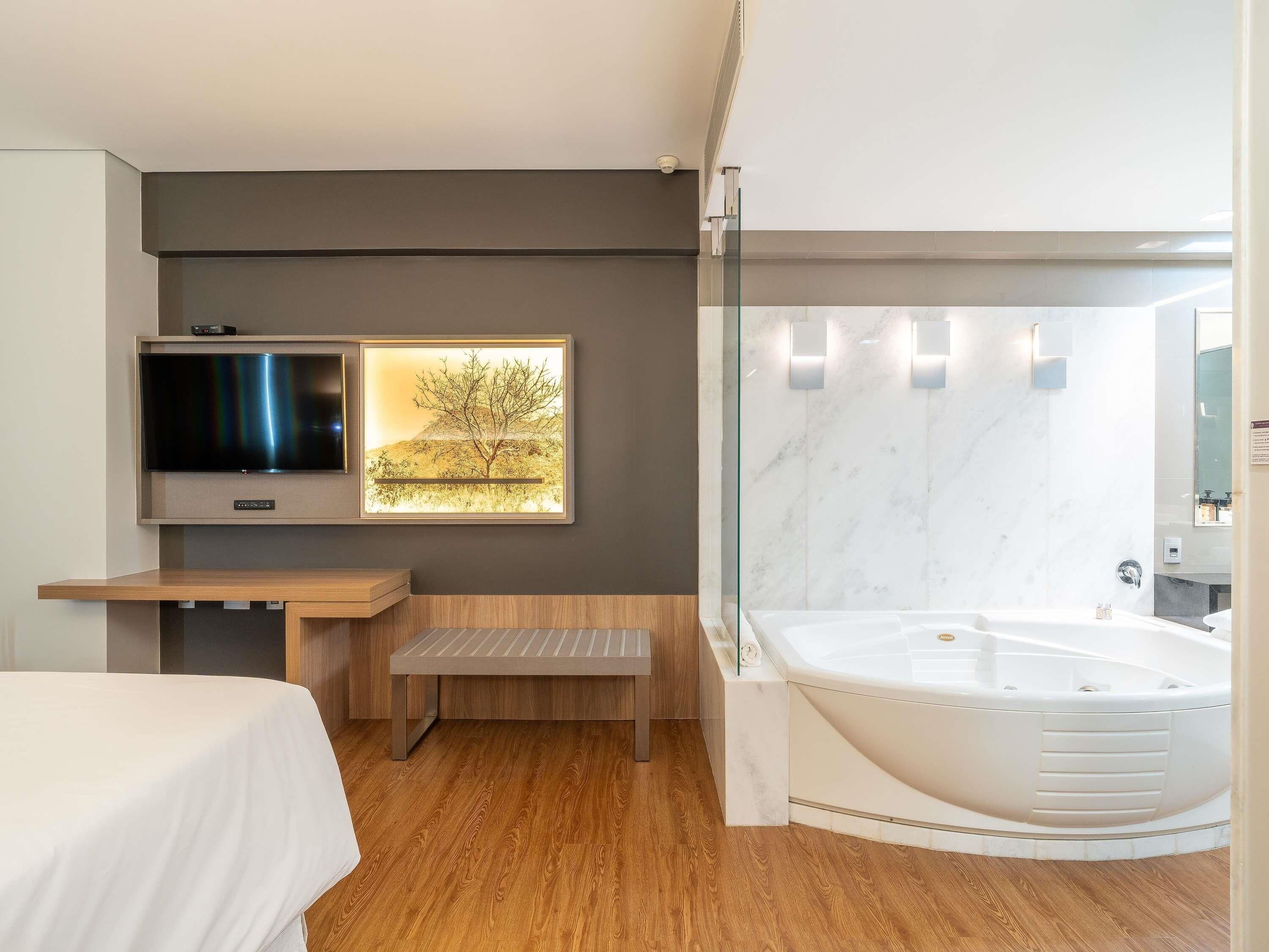A modern bathroom with a large bathtub, a wooden bench, a TV mounted on the wall, and a framed picture of a tree.
