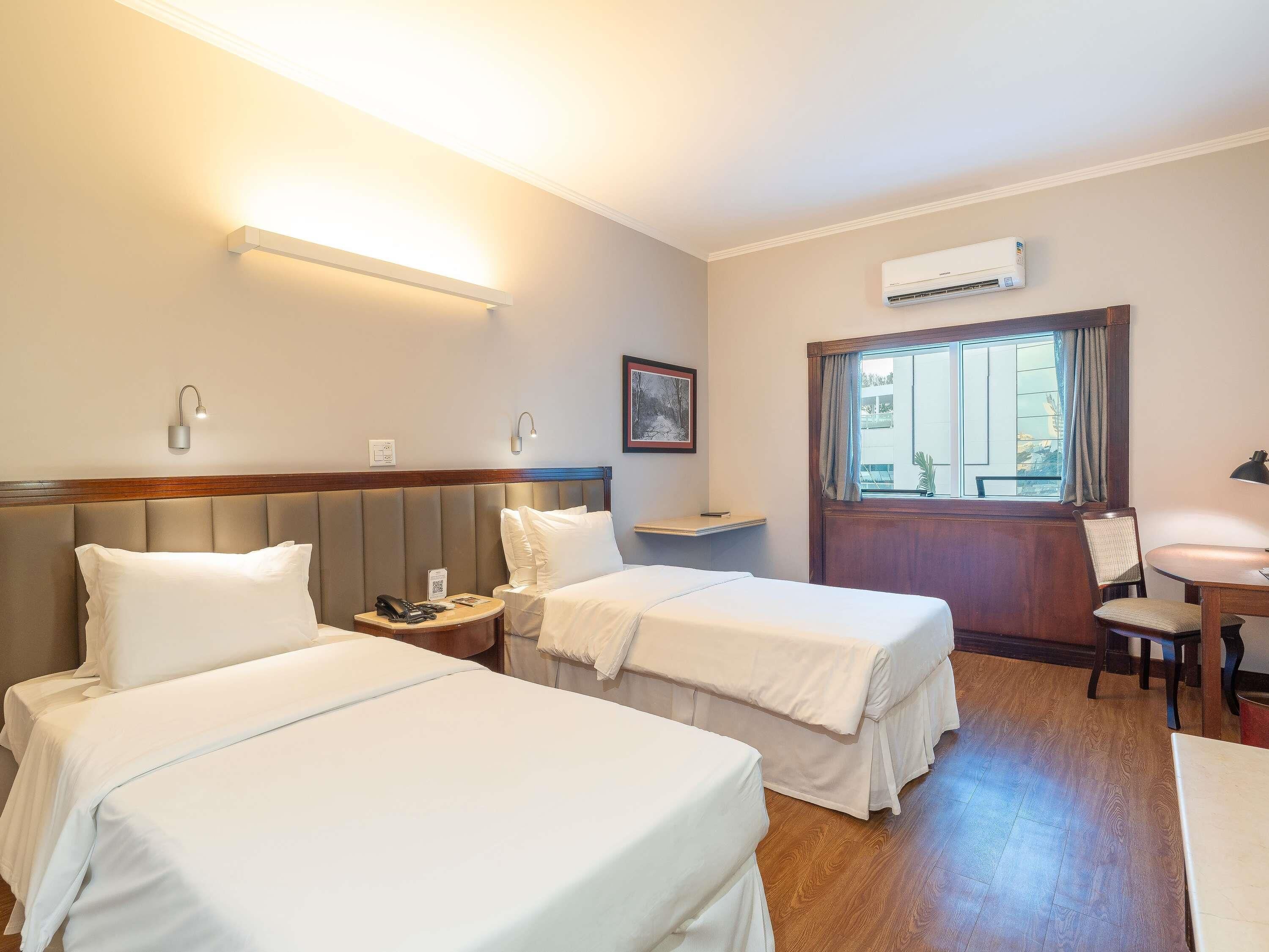A hotel room with two beds, a desk, a chair, a window with curtains, and an air conditioning unit.