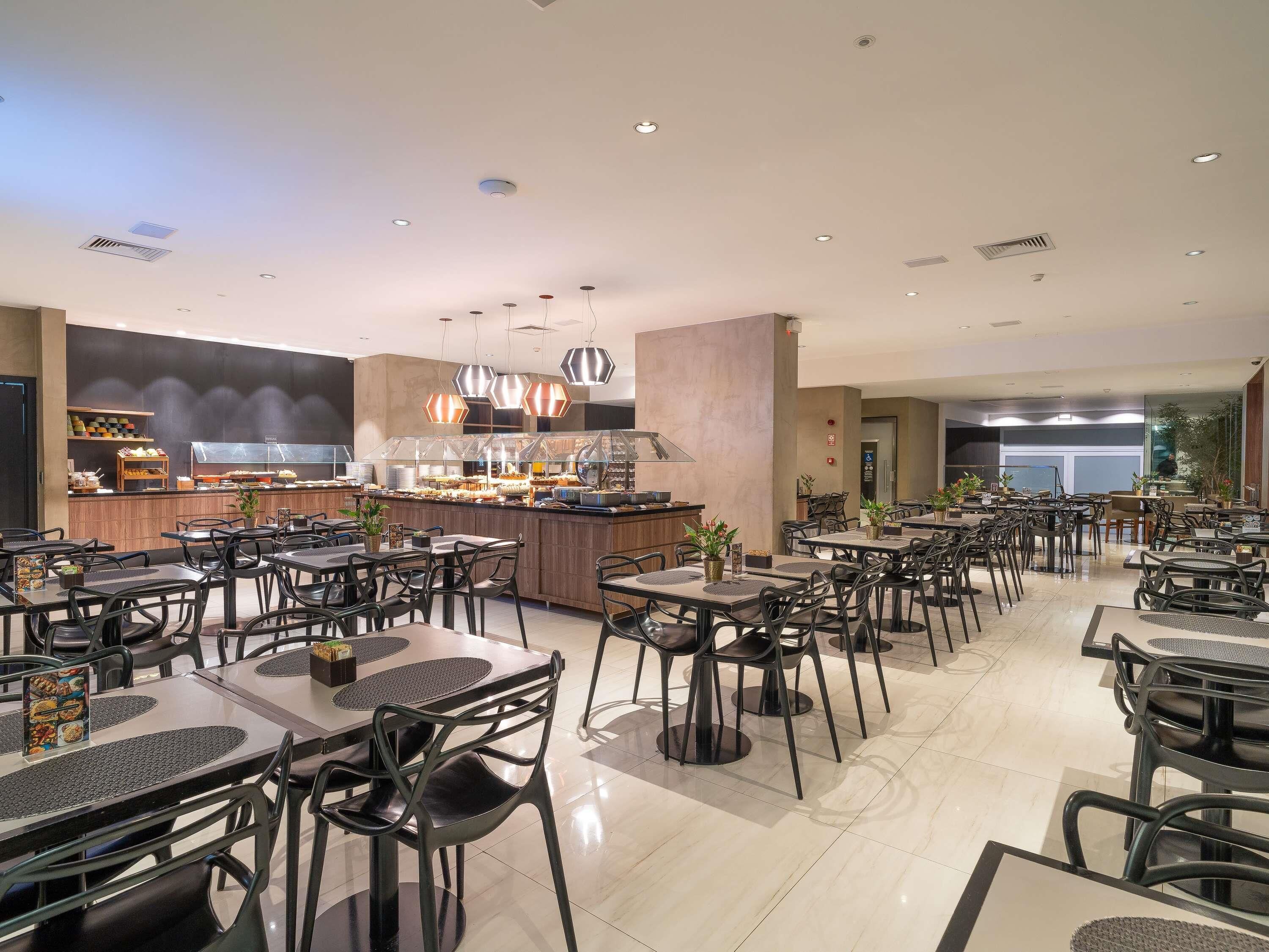 Um restaurante amplo com várias mesas e cadeiras, um balcão central com expositores de comida e luminárias no teto.