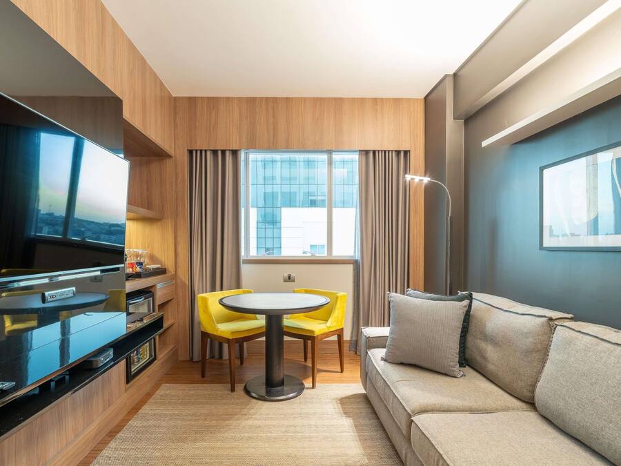 Una habitación de hotel compacta con televisor, una pequeña mesa de comedor con sillas amarillas, un sofá y un ventanal con cortinas.