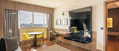 Habitación de hotel moderna con un ventanal grande, un televisor de pantalla plana, una zona de comedor y un sofá.