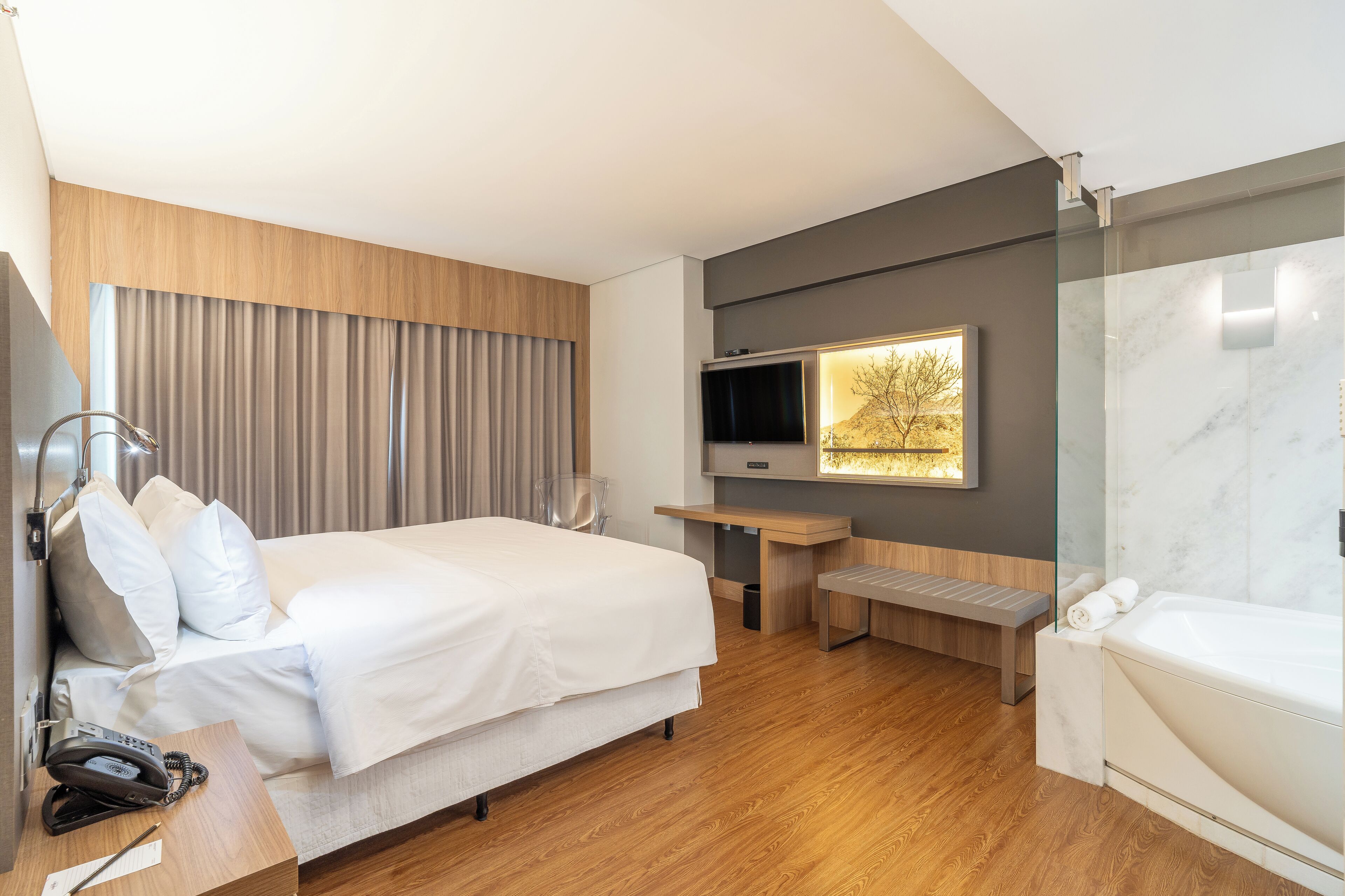 A modern hotel room with a large bed, a TV mounted on the wall, a wooden bench, and a separate bathroom area.
