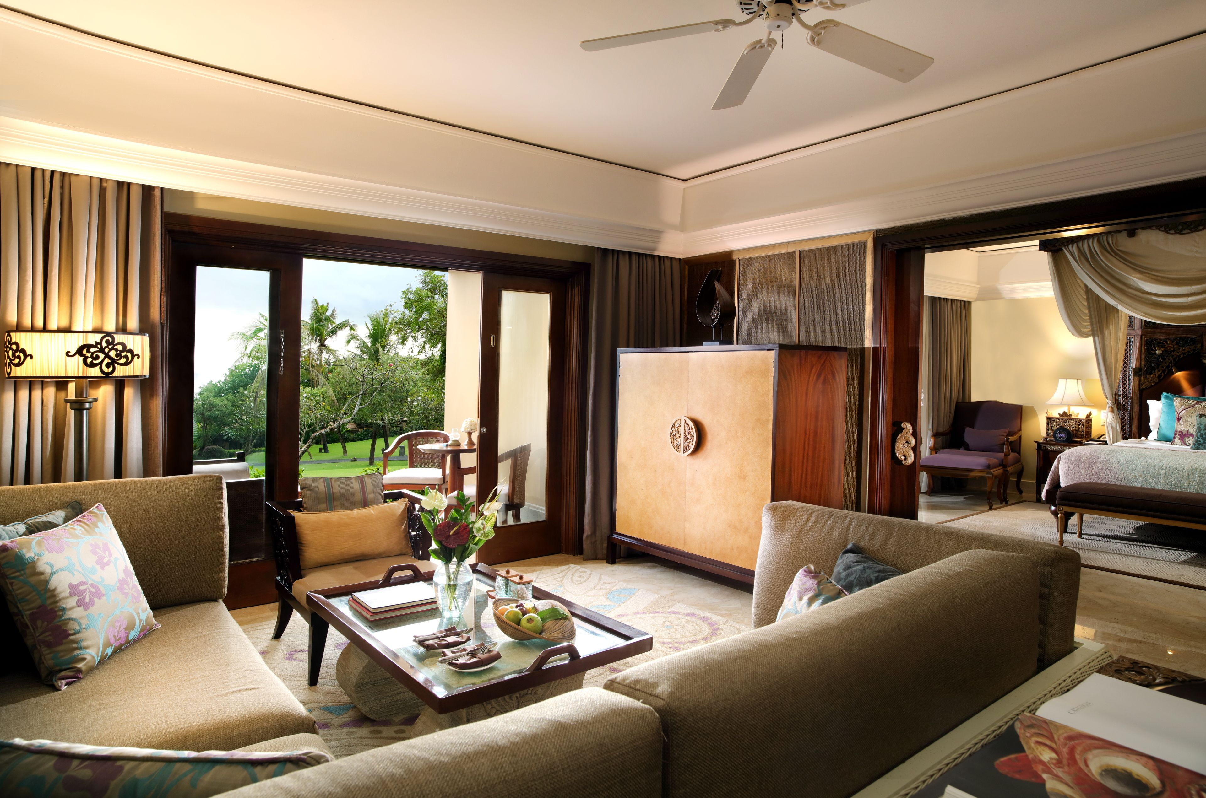 A spacious hotel room with a large bed, a sitting area with a sofa and armchairs, a dining table with a vase of flowers, and a view of the outdoors.