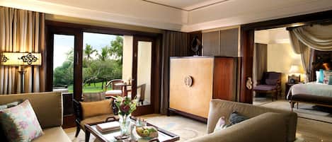 A spacious hotel room with a large bed, a sitting area with a sofa and armchairs, a dining table with a vase of flowers, and a view of the outdoors.