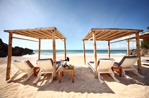 Two wooden beach cabanas with white cushioned chairs and a table with fruits and a drink. - AYANA Resort Bali (Jimbaran)