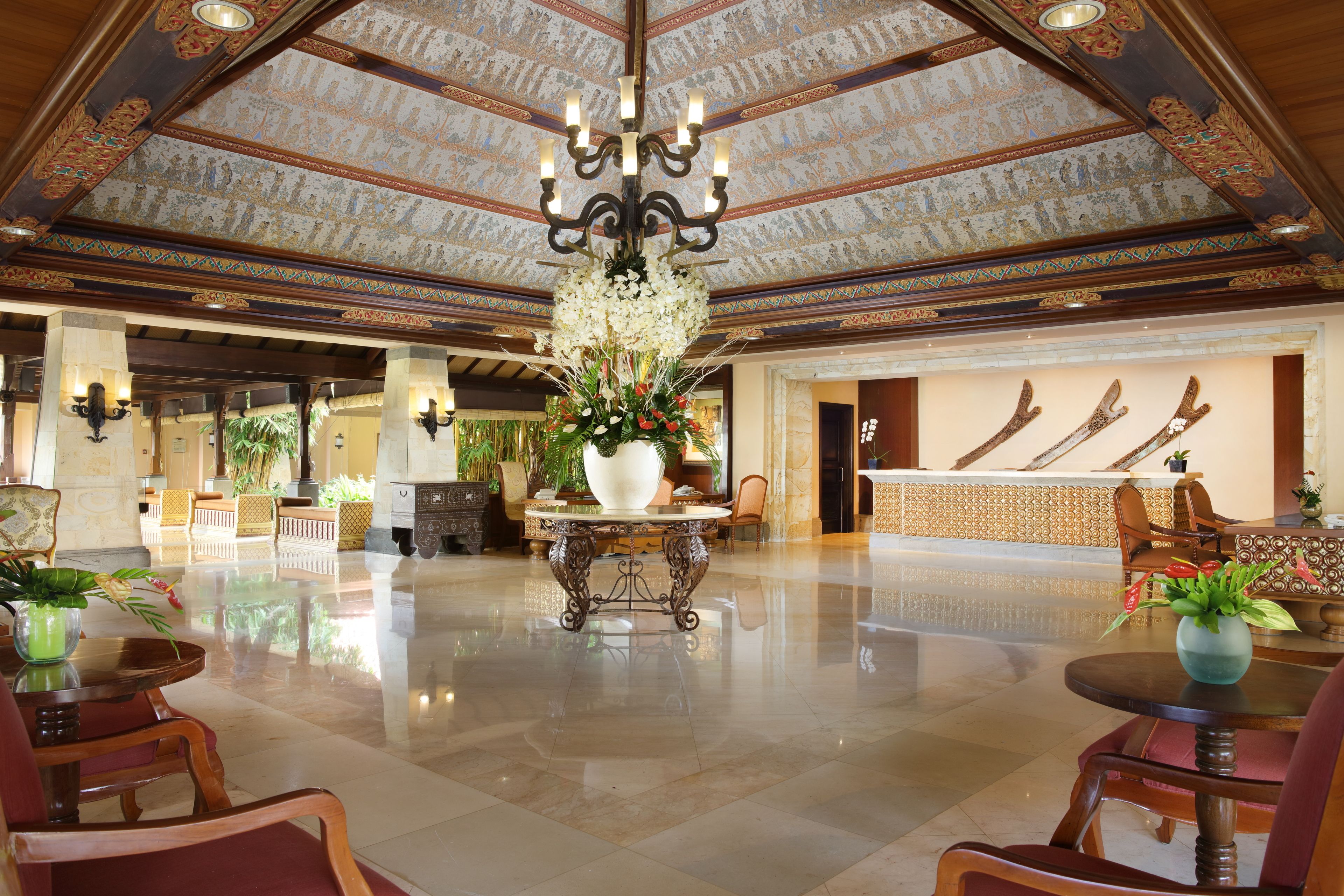 A spacious lobby with a high ceiling, ornate ceiling design, a central floral arrangement, and a reception desk with a decorative backdrop.