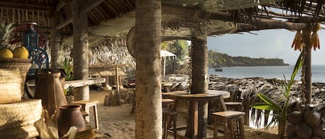 A beachside restaurant with thatched roofs, wooden tables, and chairs.