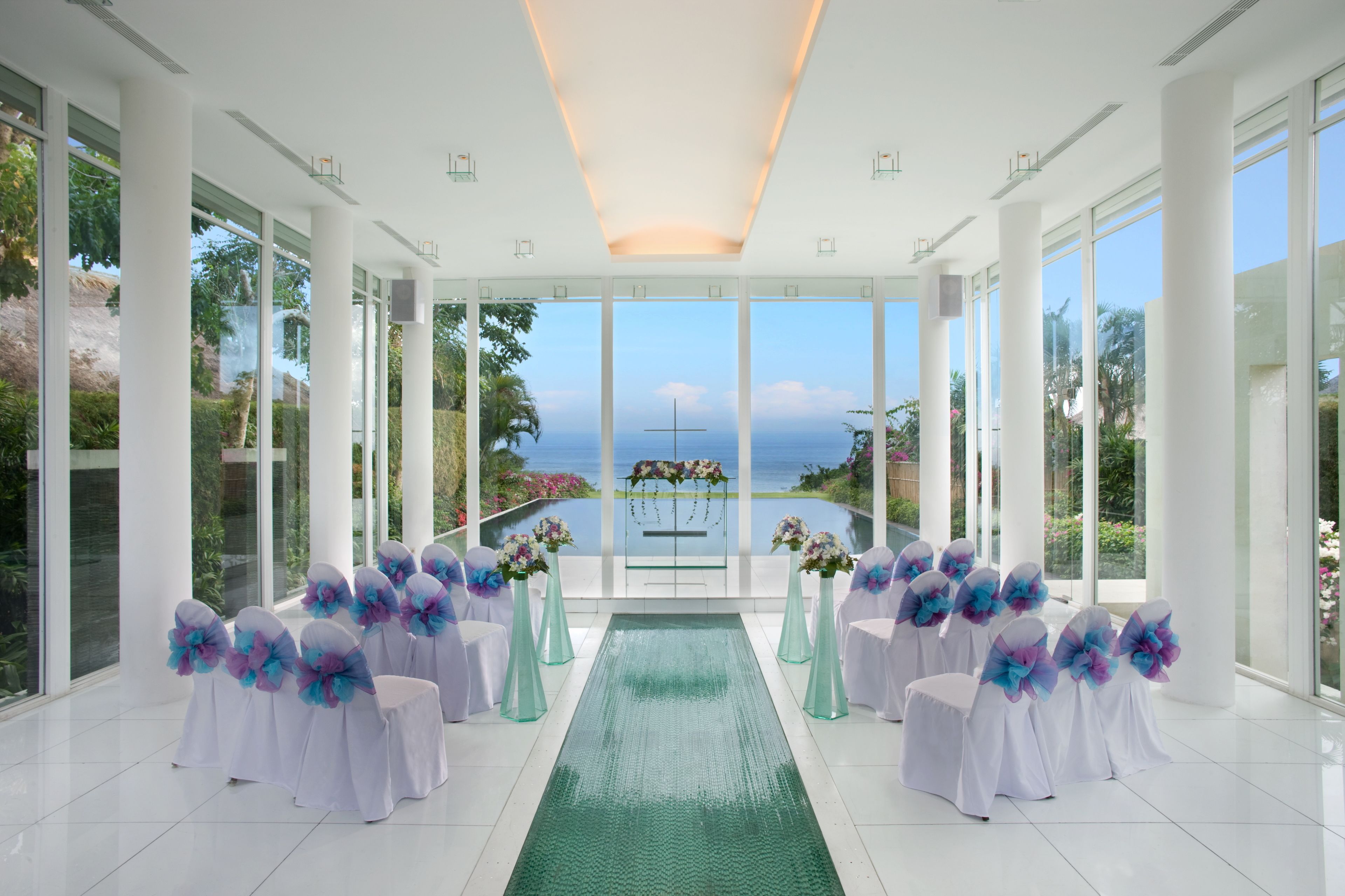 Hochzeitszeremonie mit Blick auf das Meer, weiße Stühle mit blauen und violetten Blumendekorationen und einem grünen Gangteppich.