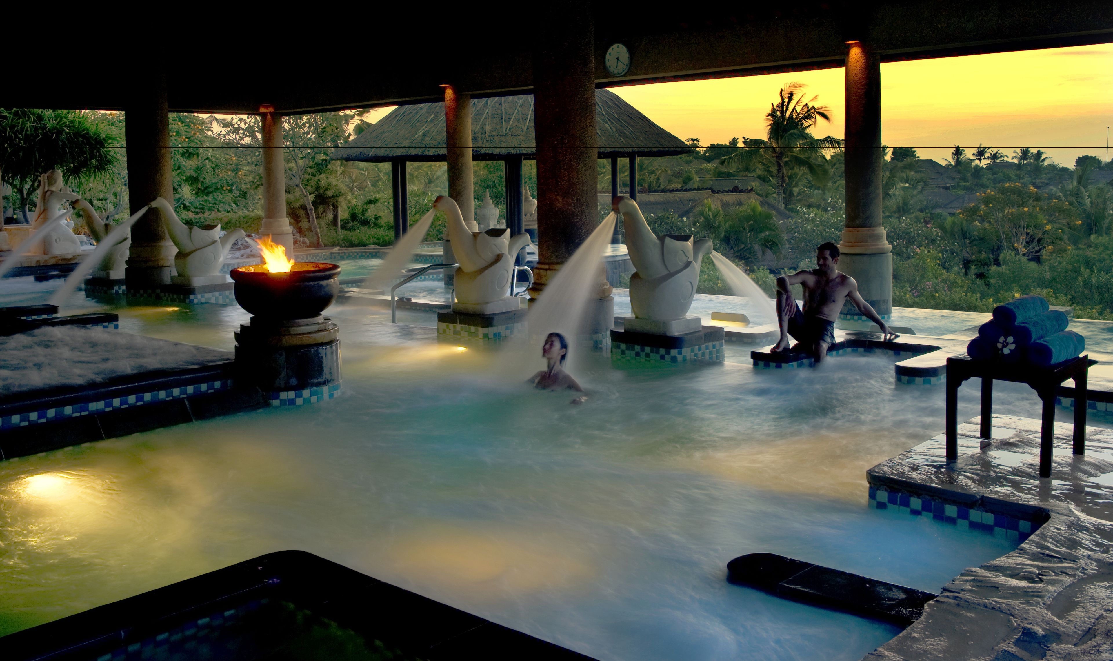 A hot spring area with a fire feature, a person relaxing, and a view of the surrounding landscape.
