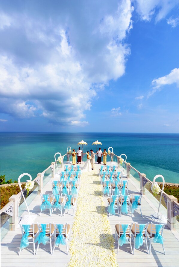 A wedding ceremony setup with chairs and a white aisle on a deck overlooking the ocean.