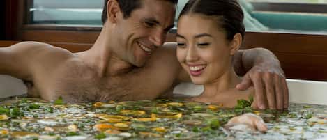 Una pareja en una bañera llena de agua y hierbas flotando.