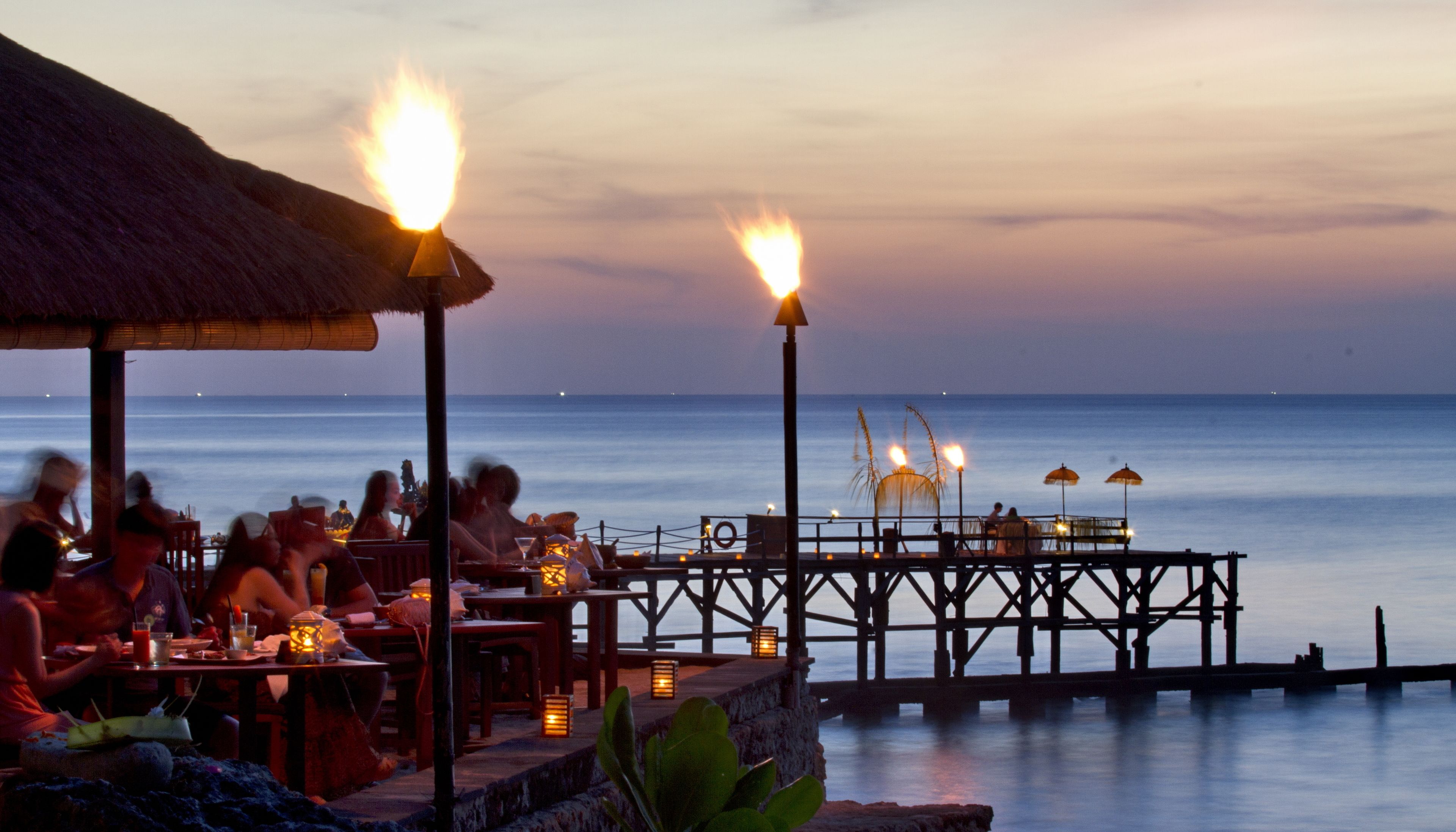 A beachside restaurant with outdoor seating overlooking the ocean, featuring thatched roofs and tiki torches.