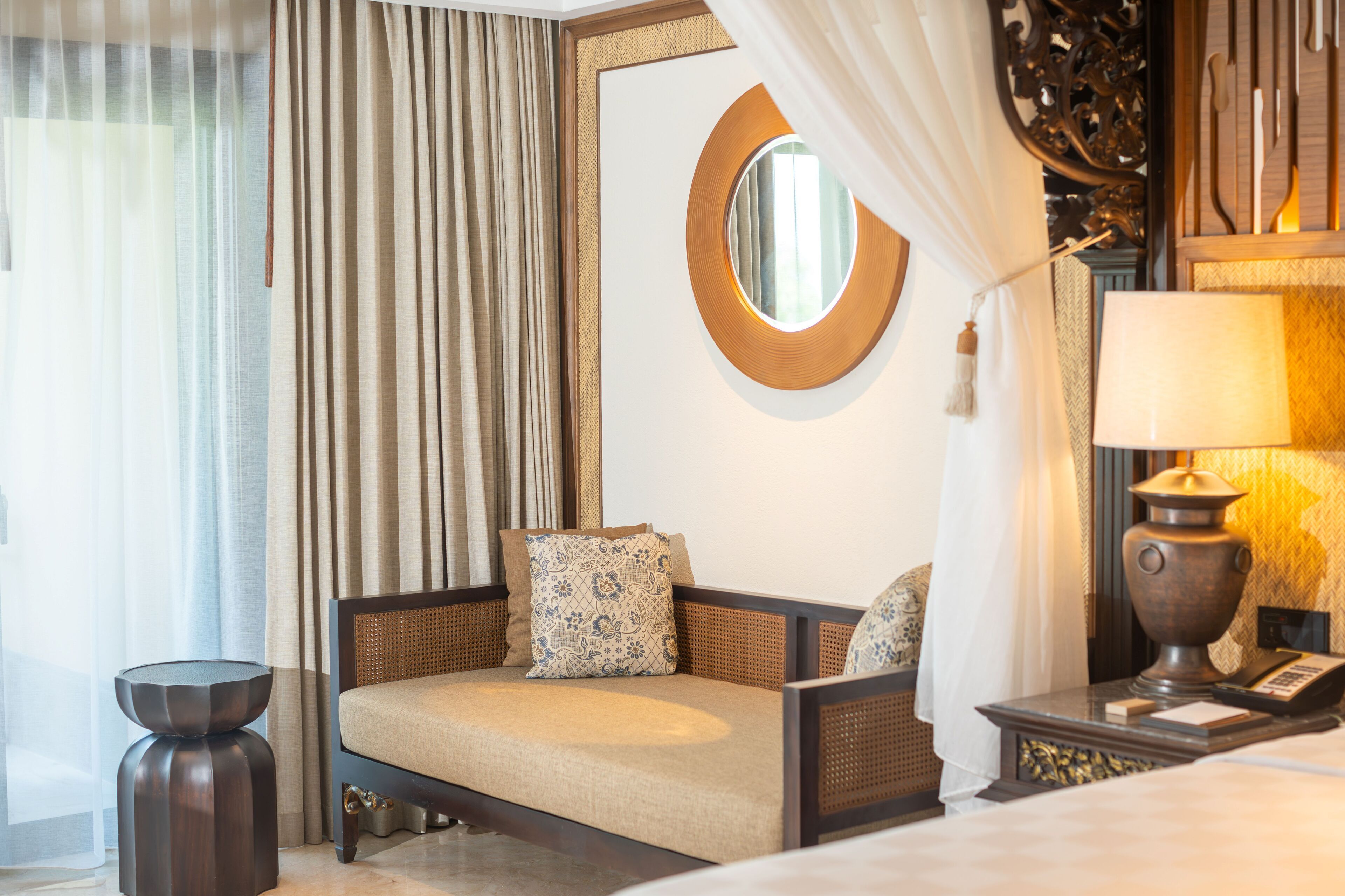 A hotel room with a sofa, a round mirror, a lamp, and a side table.