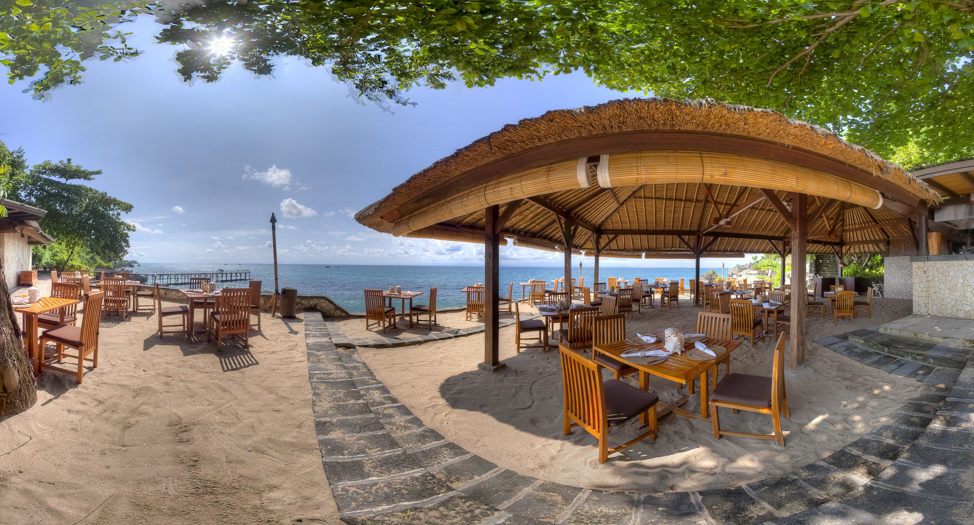 A beachside restaurant with outdoor seating, a thatched roof, and a view of the sea.