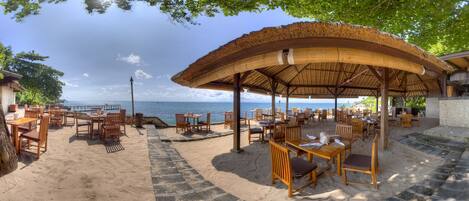 A beachside restaurant with outdoor seating, a thatched roof, and a view of the sea.