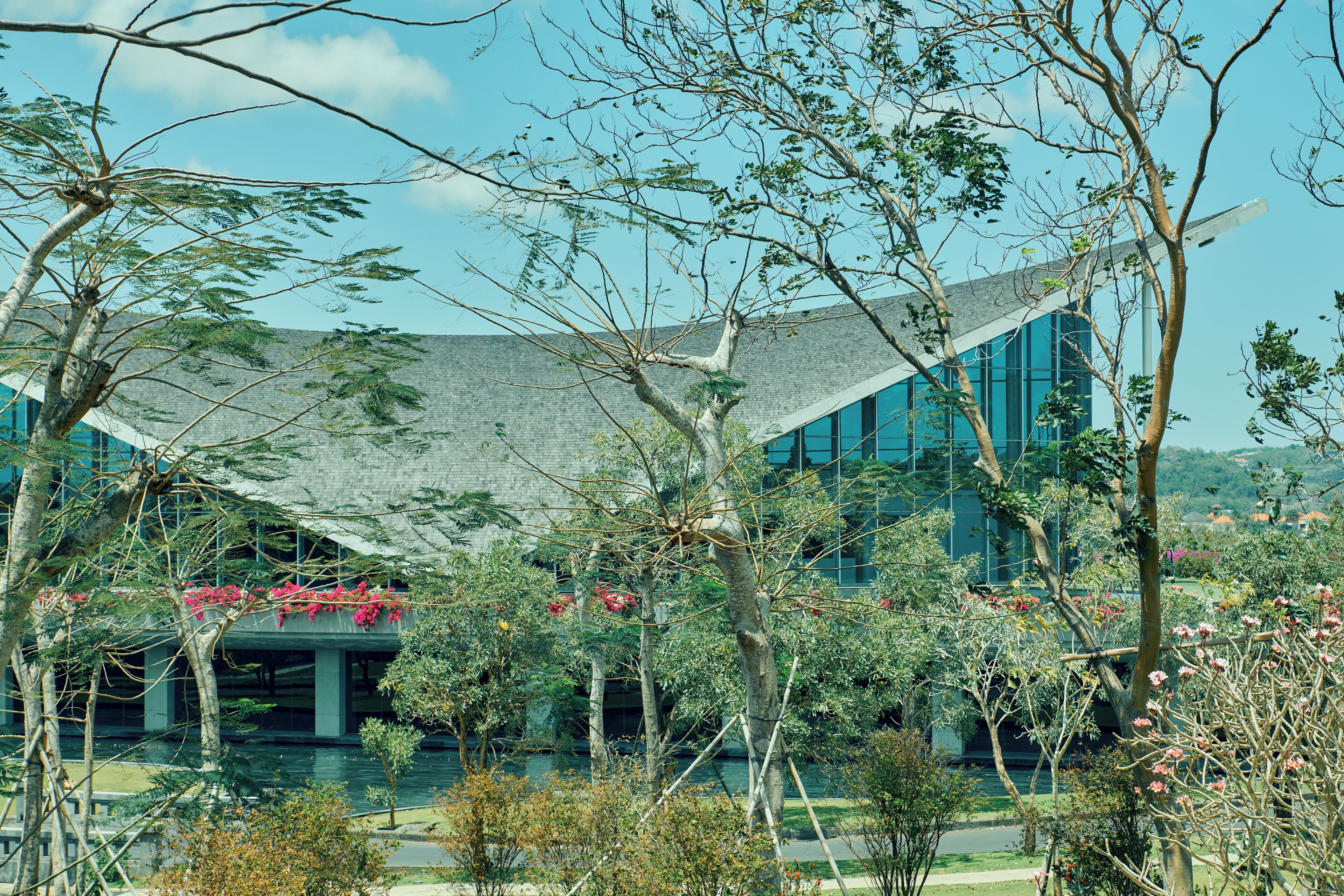 A modern building with a glass facade and a traditional roof structure, surrounded by lush greenery and flowering plants.