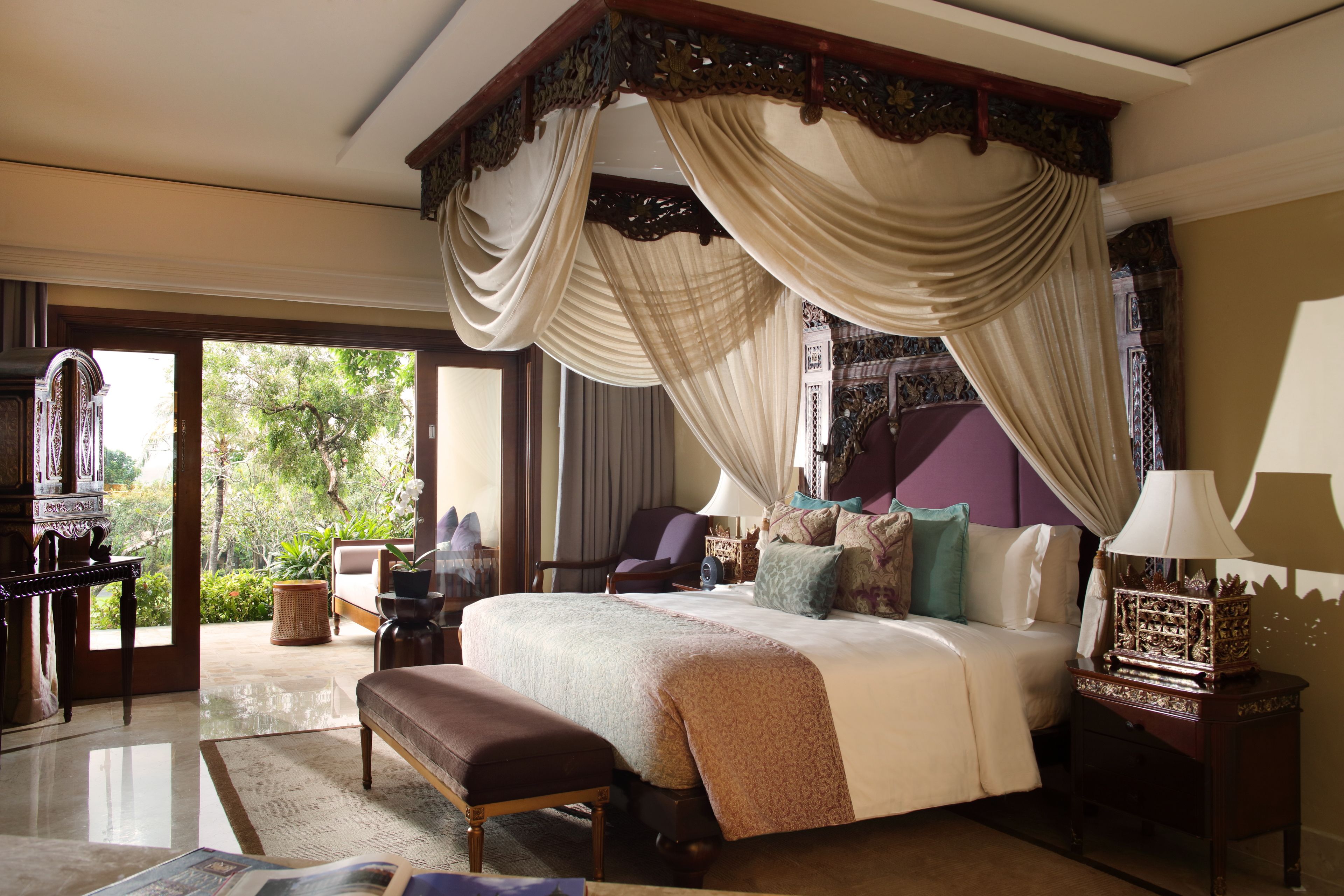 A four-poster bed with a canopy, a bench, a nightstand with a lamp, and a view of the outdoors.