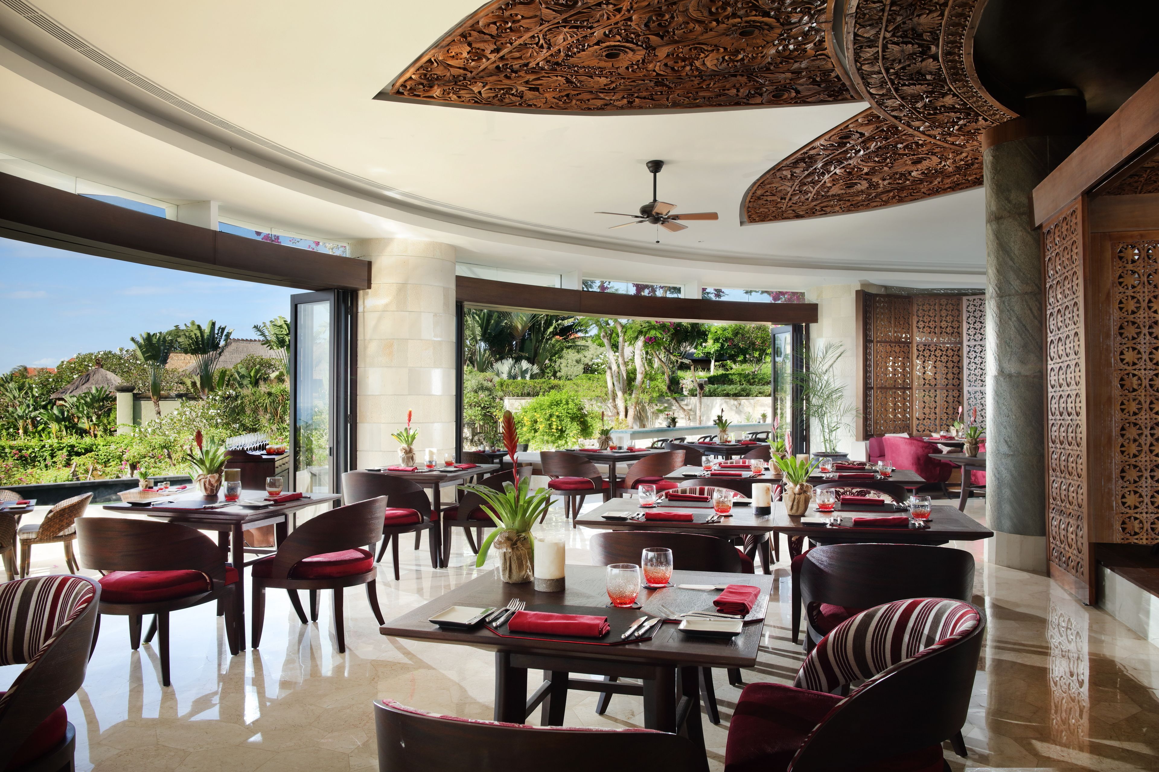 A spacious dining area with multiple tables and chairs, large windows offering a view of the outdoors, and a ceiling fan.