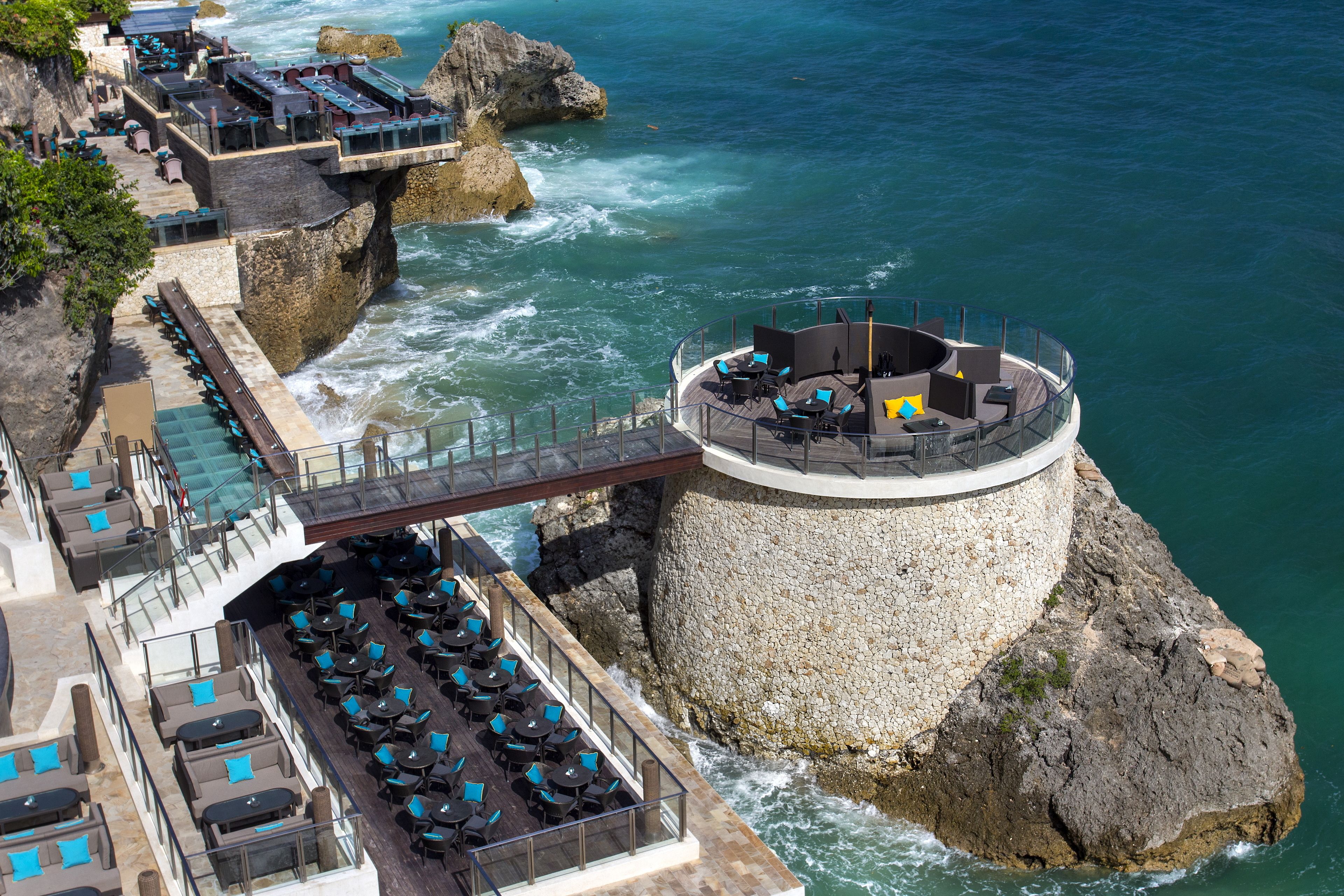 A seaside restaurant with outdoor seating overlooking the ocean, a stone structure, and a bridge connecting to the main building.