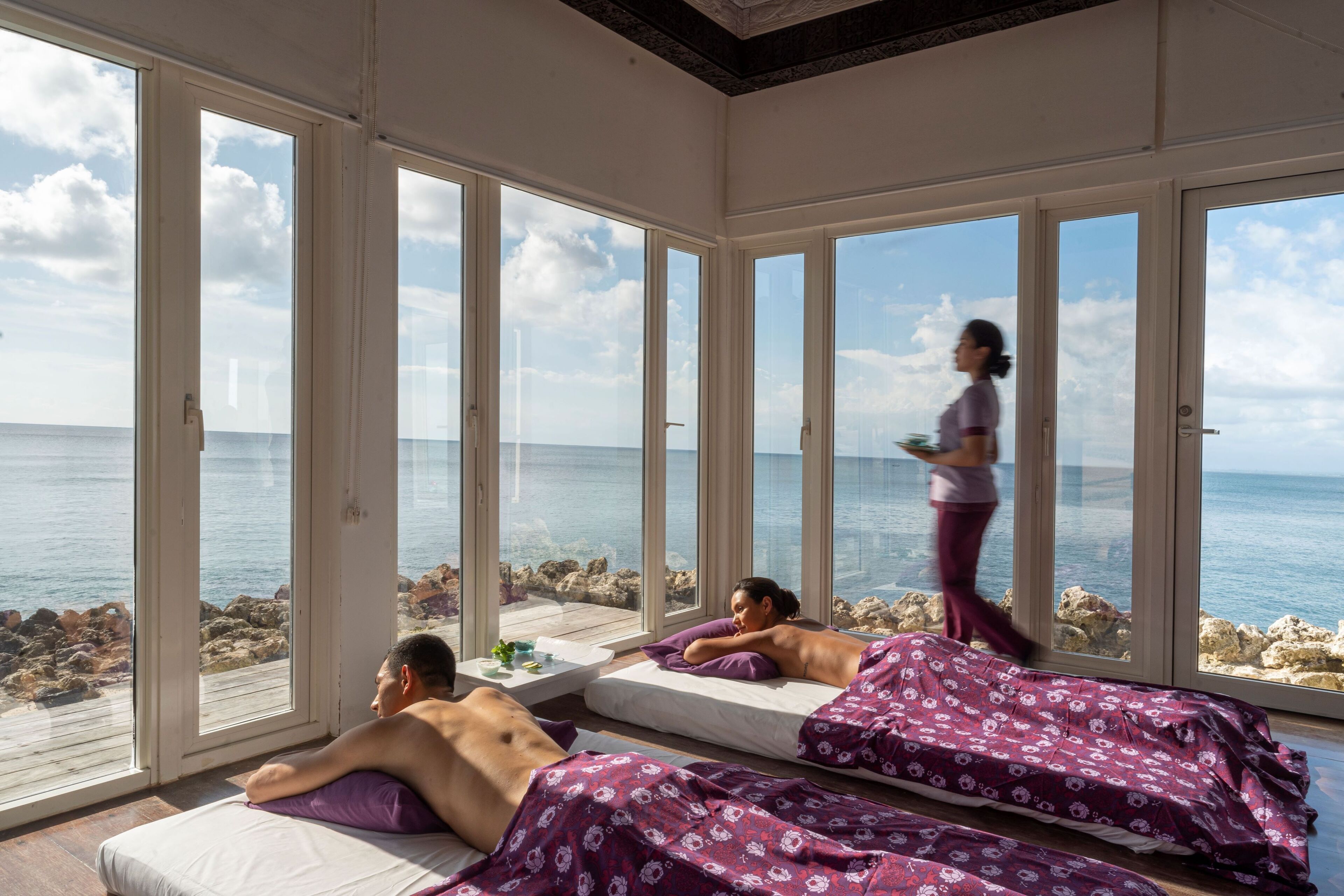 Una habitación con amplios ventanales con vistas al mar, dos camas con ropa de cama estampada de flores moradas, y una persona caminando.