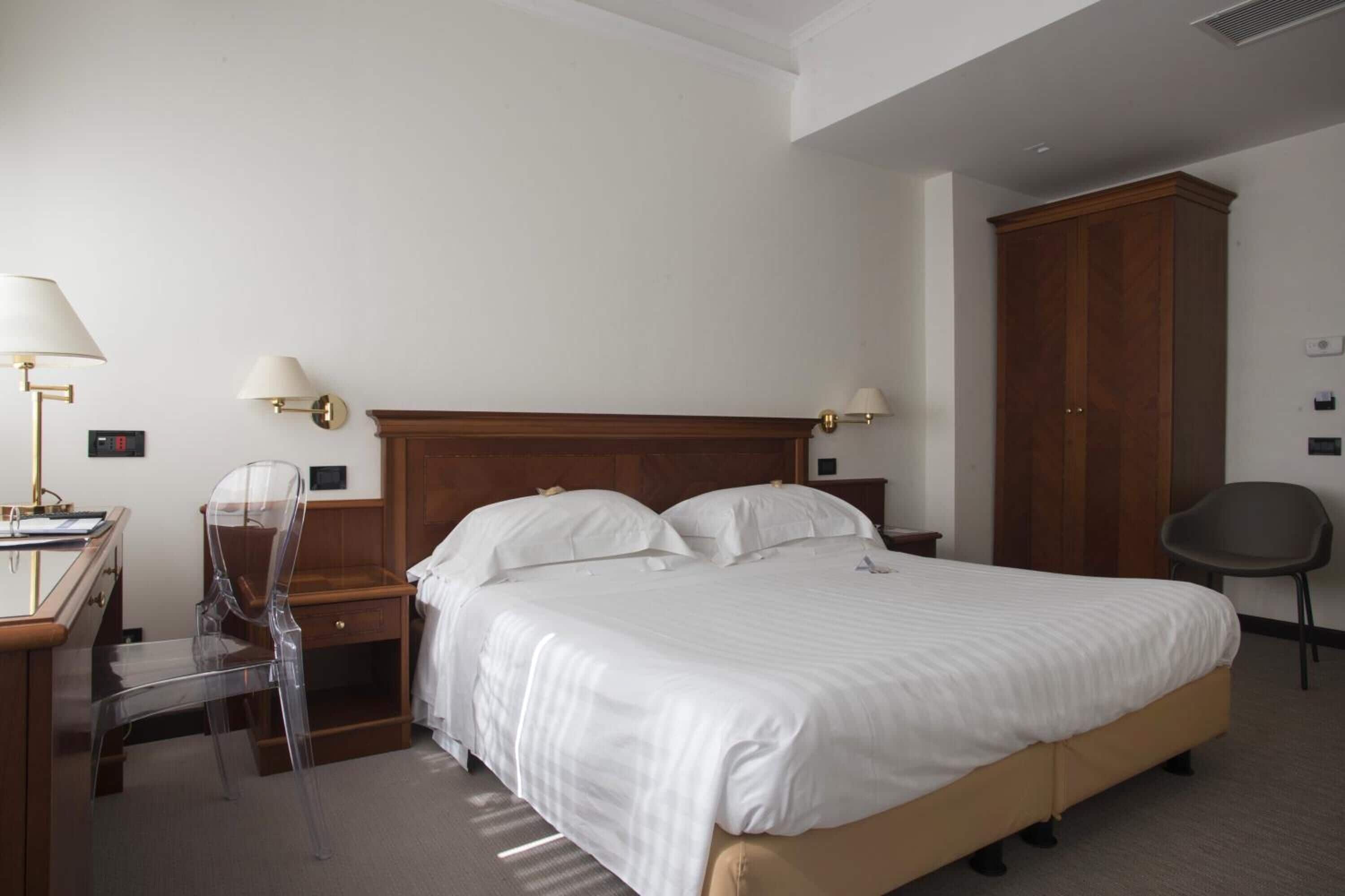Habitación de hotel con una cama grande, un escritorio con silla, dos lámparas de pared, un armario y una silla.