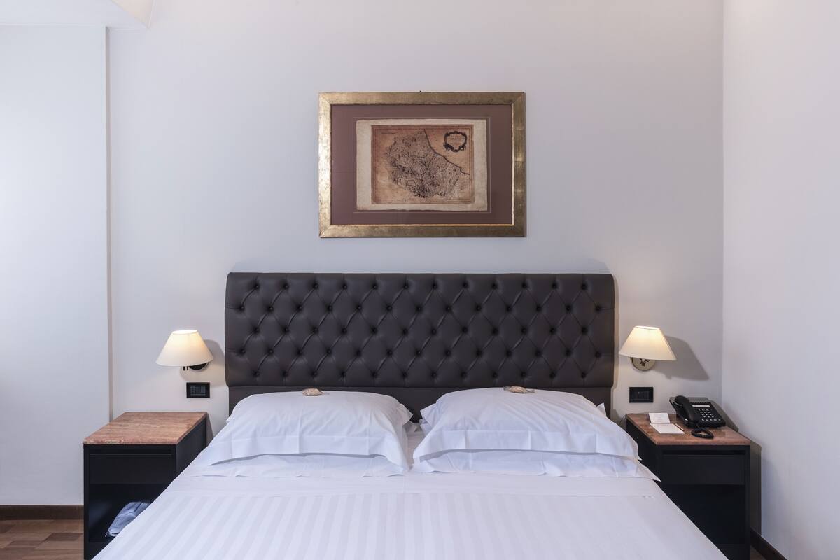A neatly made bed with white linens, a dark tufted headboard, and two bedside tables with lamps and a phone.