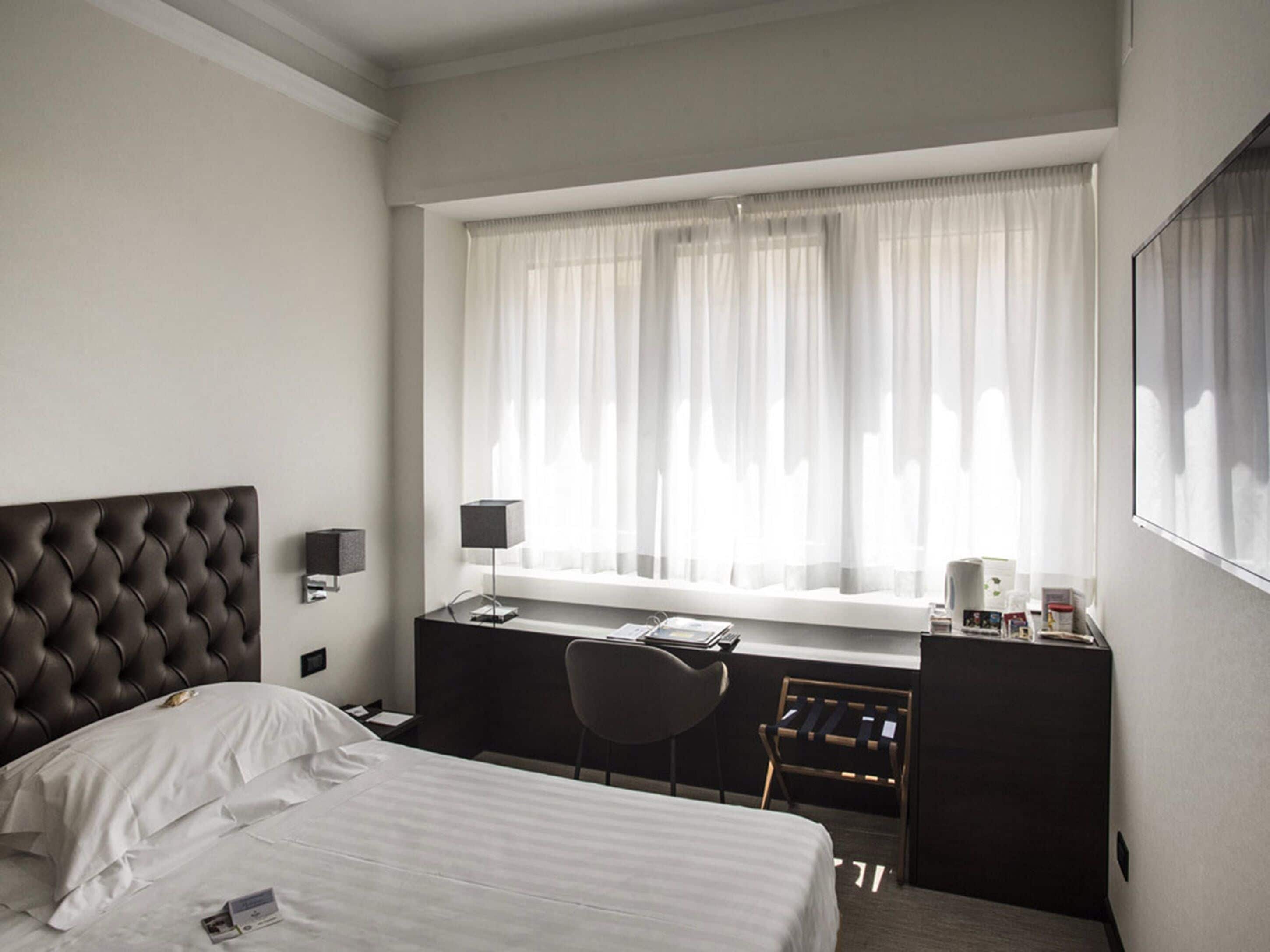 Una habitación de hotel con cama, escritorio, silla y ventana con cortinas.