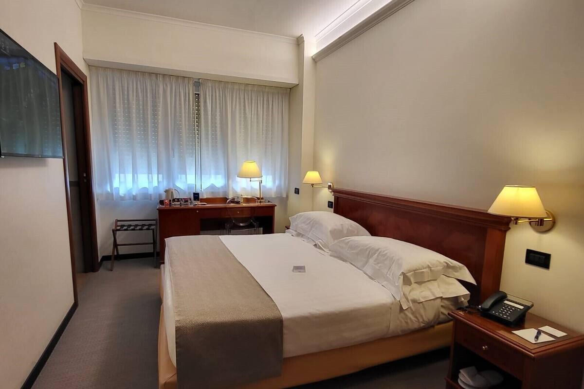 A hotel room with a bed, two bedside lamps, a desk with a lamp, a chair, and a telephone on a nightstand.