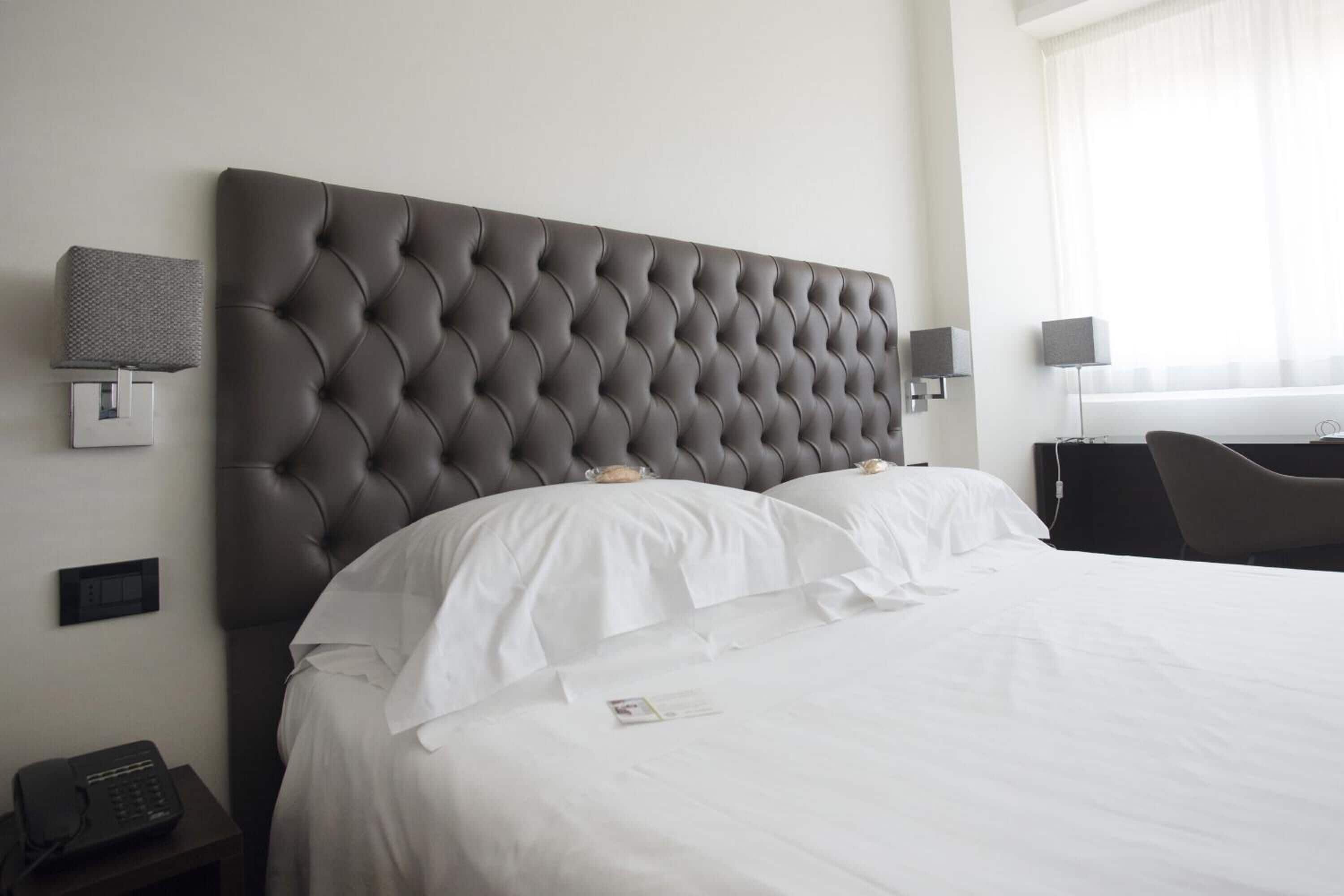 Una habitación de hotel con cabecera acolchada, ropa de cama blanca, una mesita de noche con teléfono y una lámpara.