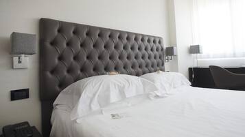 Una habitación de hotel con cabecera acolchada, ropa de cama blanca, una mesita de noche con teléfono y una lámpara.