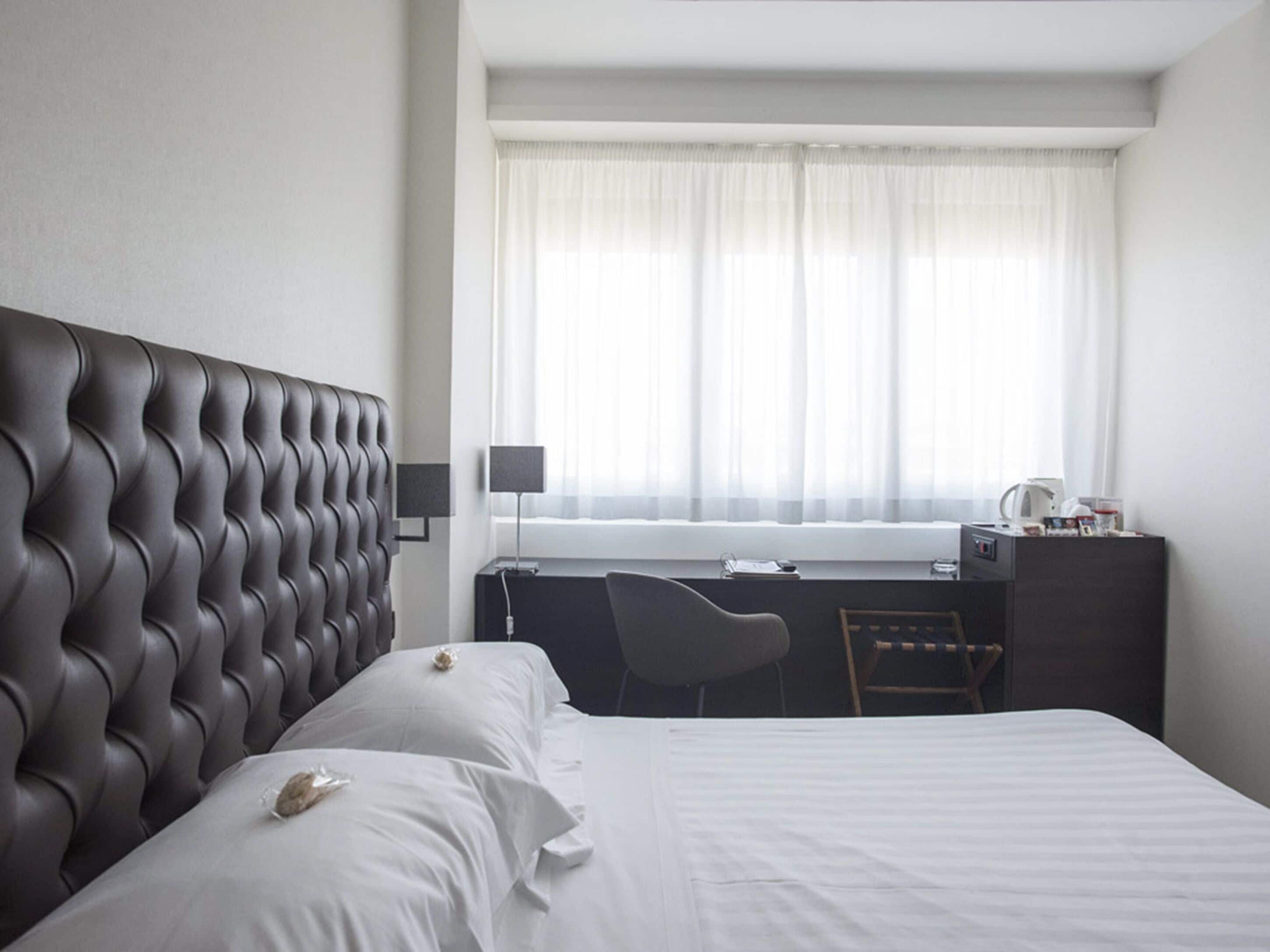 Una habitación de hotel con cabecera capitonada, cama, escritorio con silla, ventana con cortinas translúcidas y cafetera sobre el escritorio.
