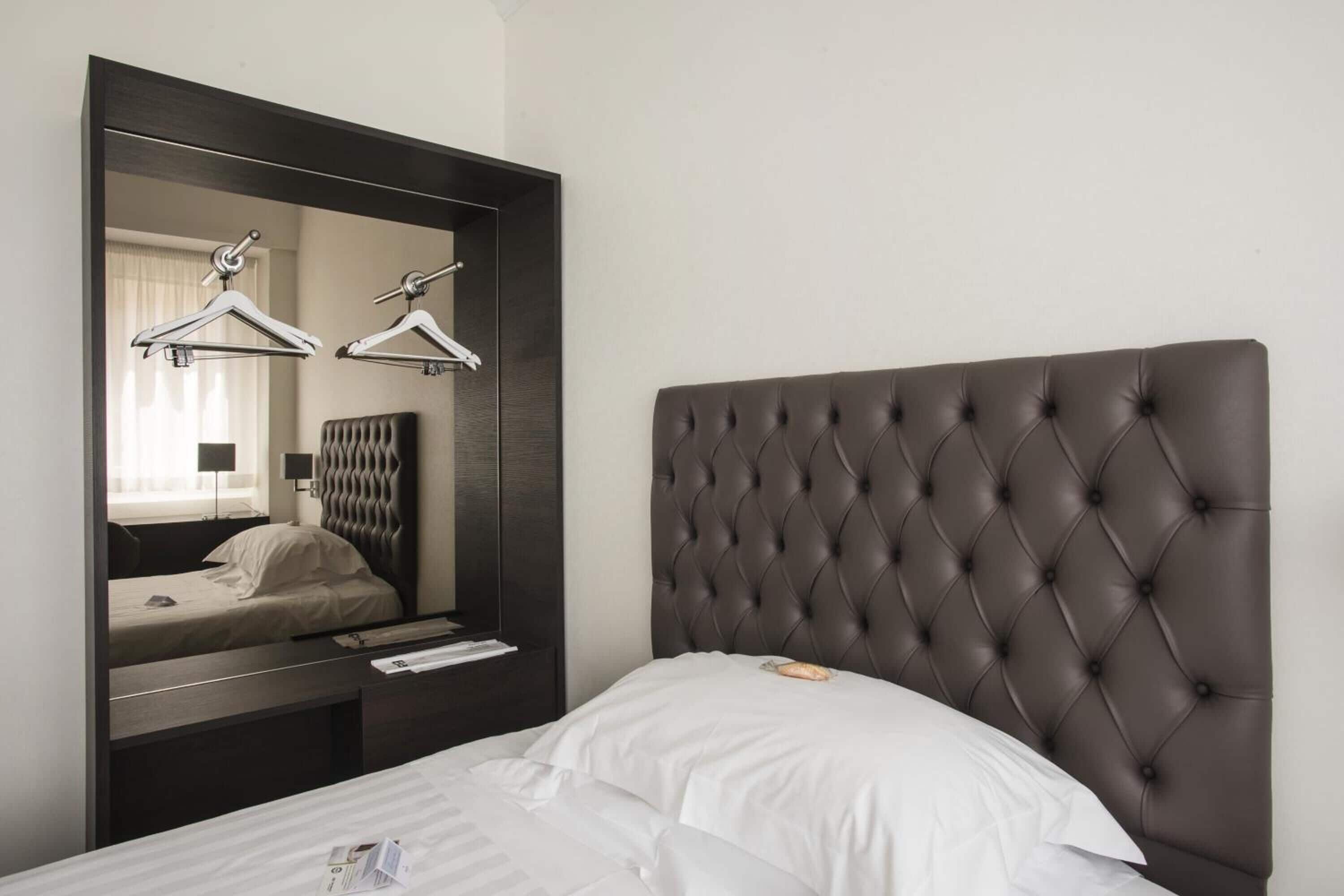 A hotel room with a large mirror, a tufted headboard, a bed with white linens, and a nightstand with a lamp and a book.