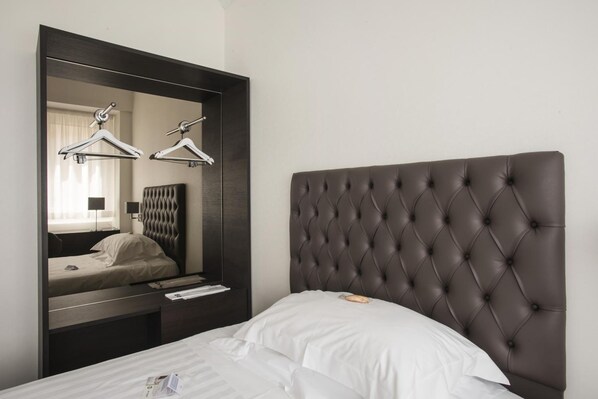 A hotel room with a large mirror, a tufted headboard, a bed with white linens, and a nightstand with a lamp and a book.