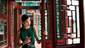 Une femme vêtue d’une robe verte, tenant un verre à martini, se tient dans un couloir aux portes rouges et aux encadrements de fenêtres verts.