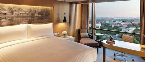 A hotel room with a large bed, a desk with a chair, a small table, and a view of the city through large windows.