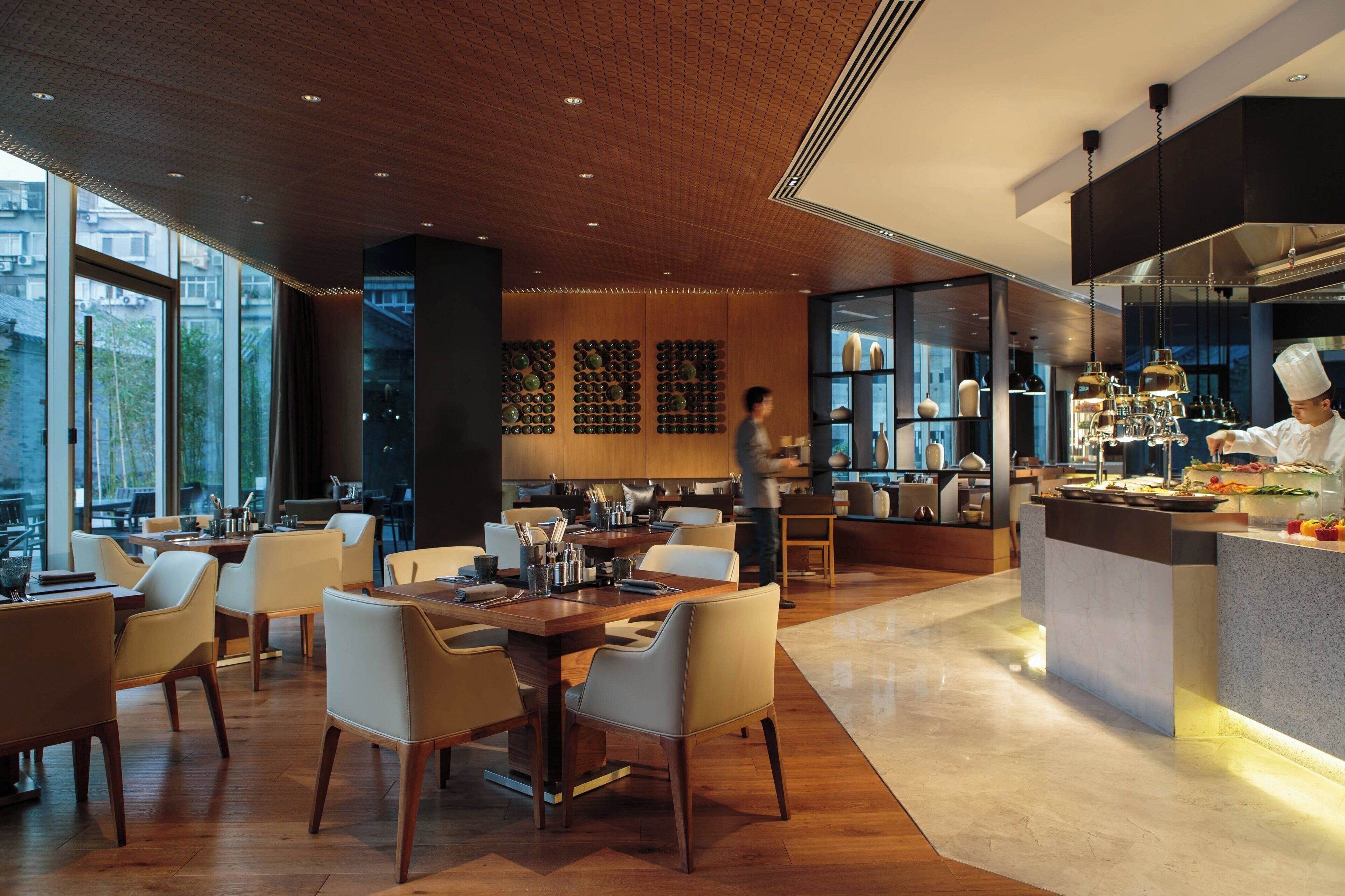 A modern restaurant with wooden flooring, a central dining area with tables and chairs, a kitchen area with a chef, and large windows providing natural light.