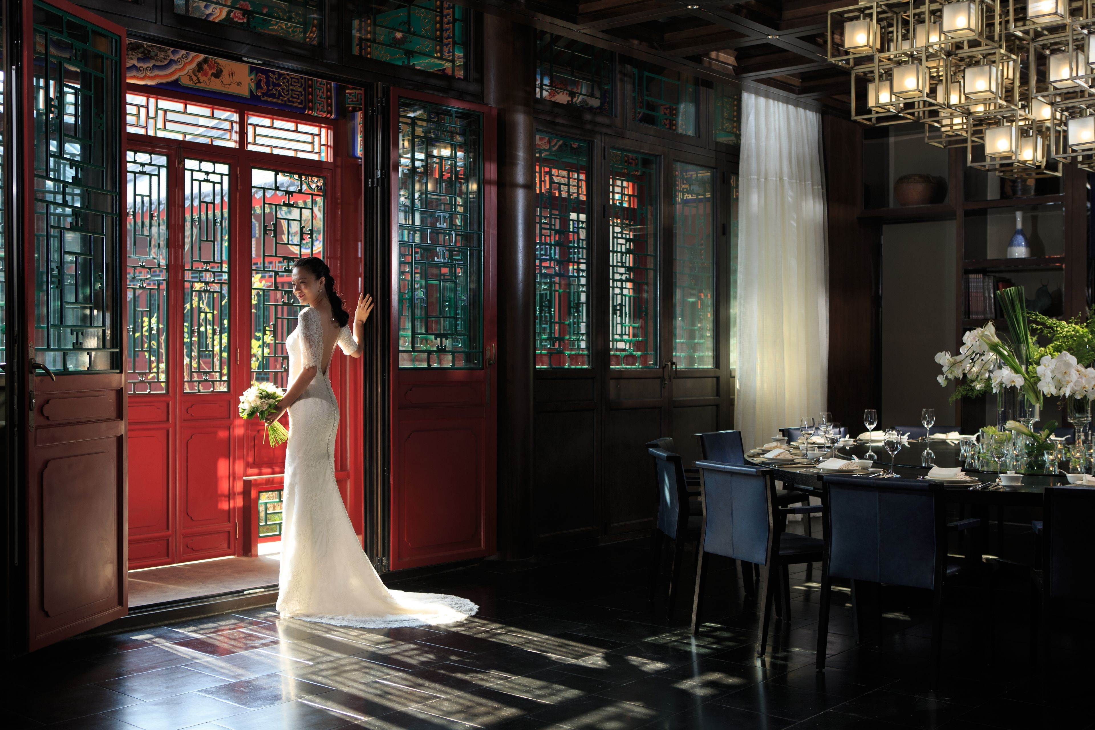 A bride in a white dress standing in a room with traditional Chinese doors and a dining area set up for a wedding reception.